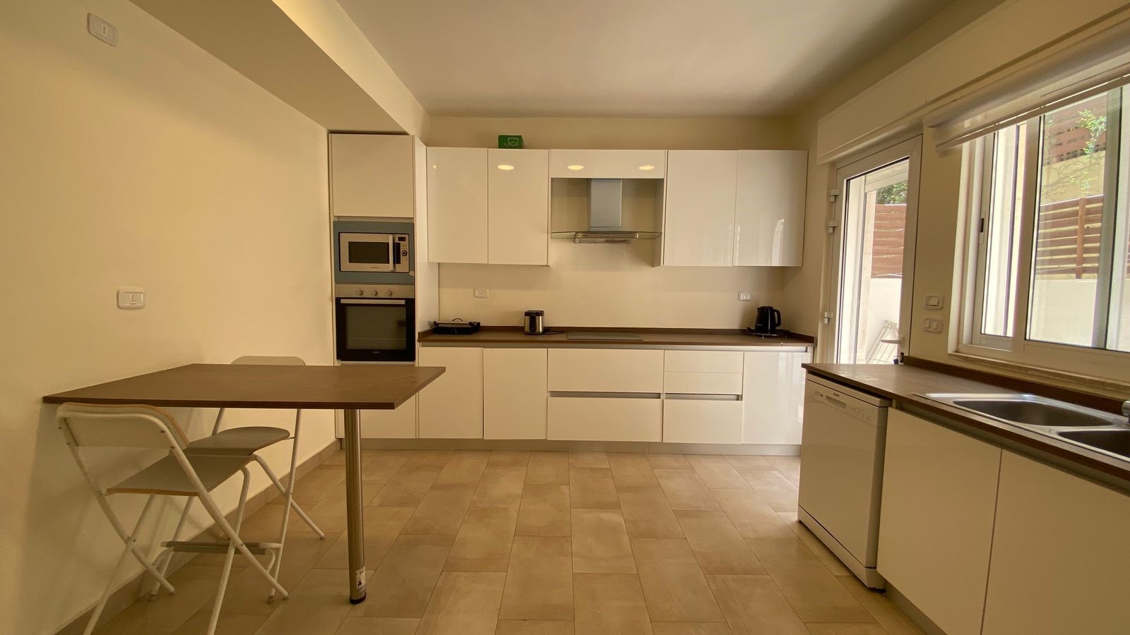 90740691-de1b-475e-a847-e0c14cddf2a6 Modern kitchen with white high-gloss cabinets, brown counters, built-in microwave and oven, and a small dining table with two folding chairs on the left.