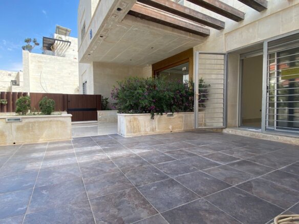 Modern patio with large dark tiled floor, glass sliding doors, and flowering shrubs in planter boxes outside a beige stone house.