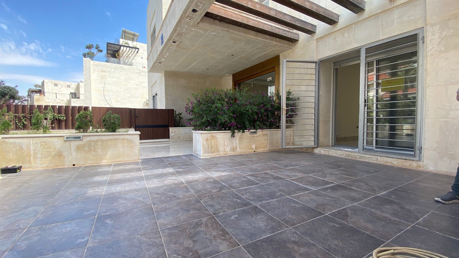 908b76de-6e5f-4269-884c-17023d8e549e Modern patio with large dark tiled floor, glass sliding doors, and flowering shrubs in planter boxes outside a beige stone house.