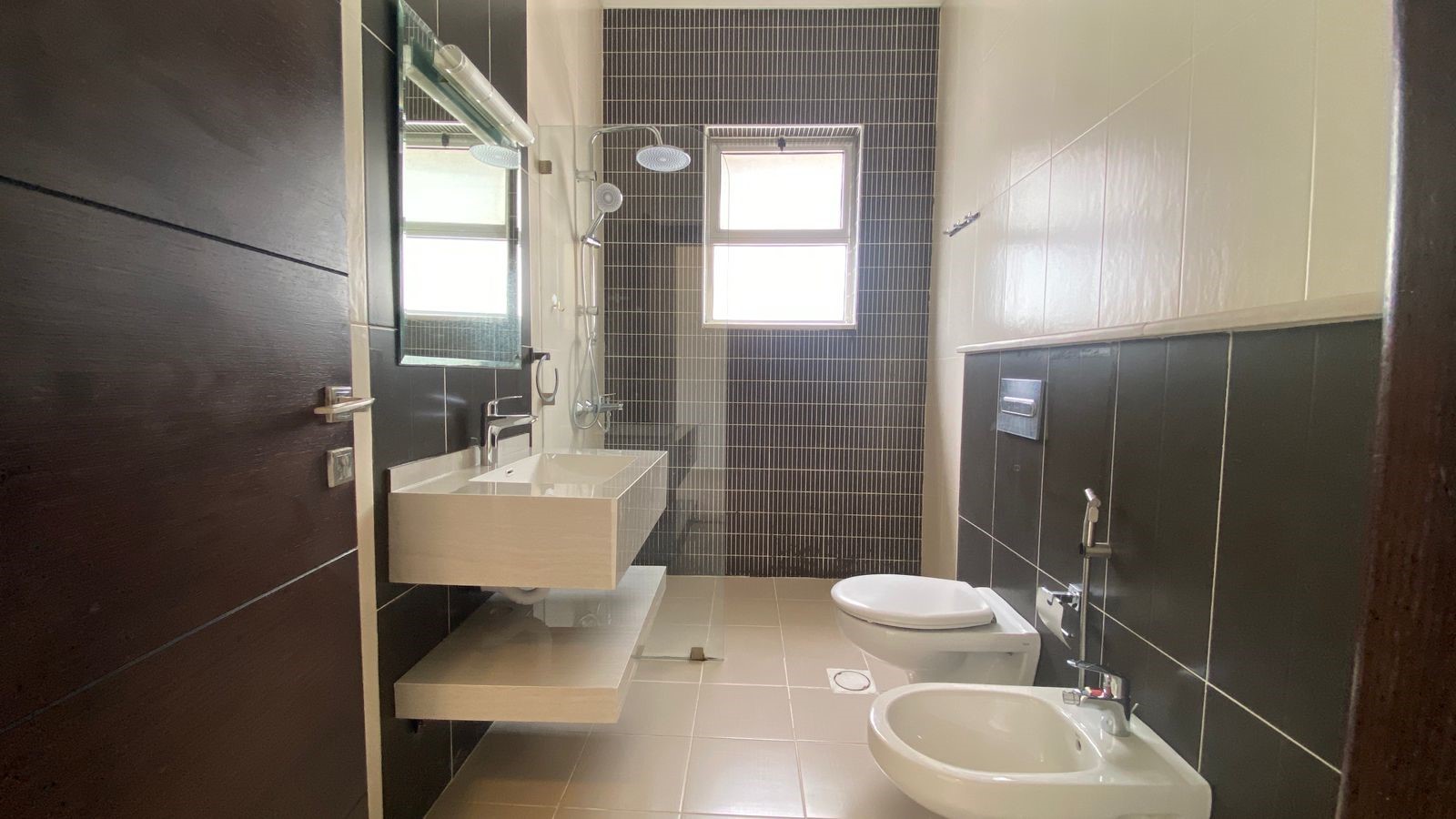 910e46af-1e1b-4278-bb56-7847935367bb Modern bathroom with dark tile accent wall, white floating sink, and glass-enclosed shower across from a toilet and bidet. Behind is a small window.
