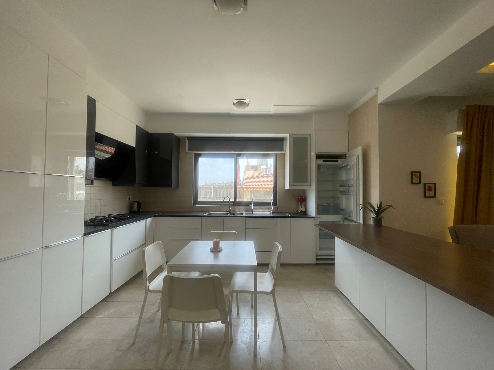 92130979-711c-461c-a97d-0445e193632d Contemporary white kitchen with glossy cabinets, a black range hood, gray counters, and a white dining table with four chairs in the center.