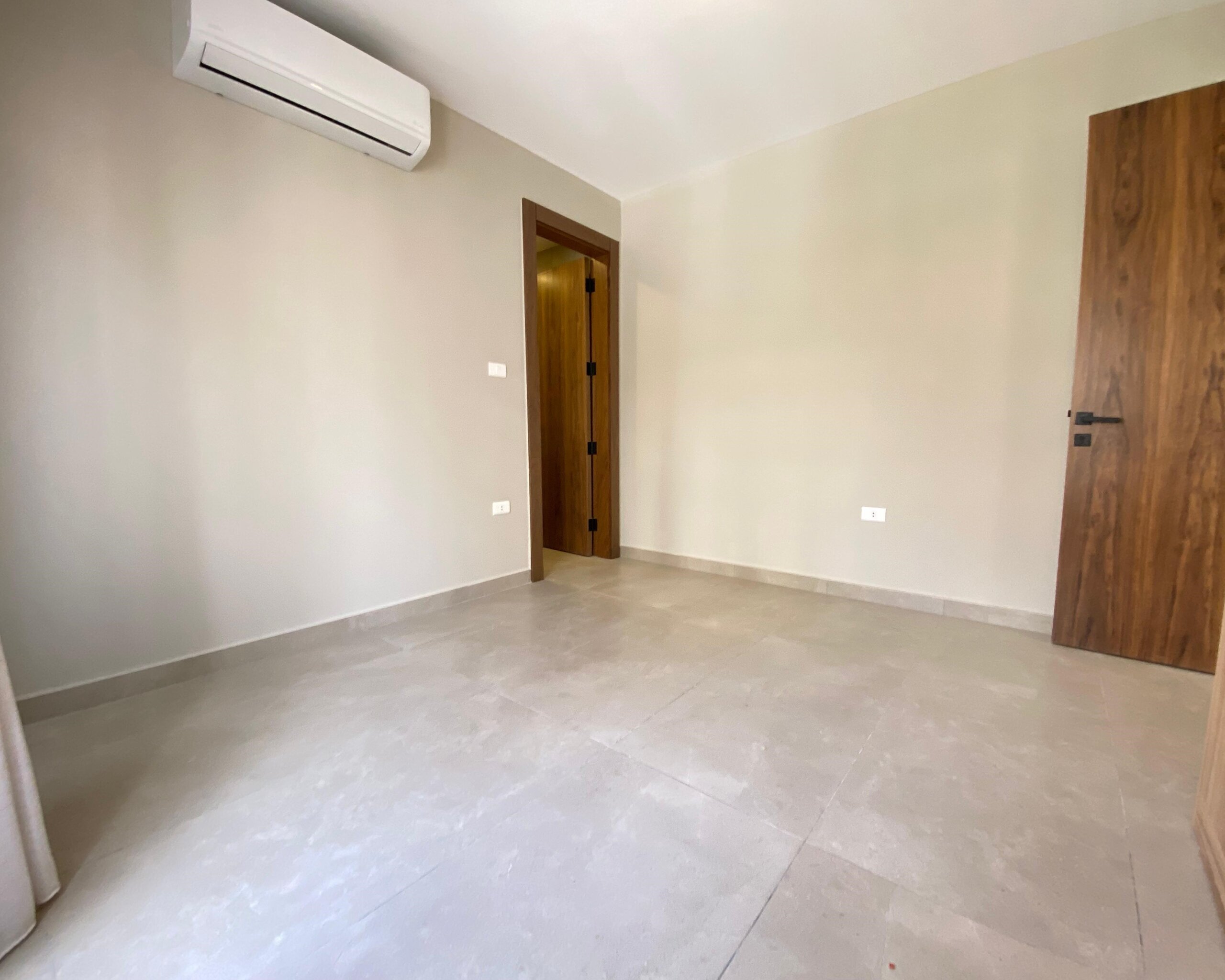93366d5c-af03-4da4-997d-bcdd77086bf5 Vacant empty room with beige walls, large tiled floor, wall-mounted white air conditioner, and two wooden doors.
