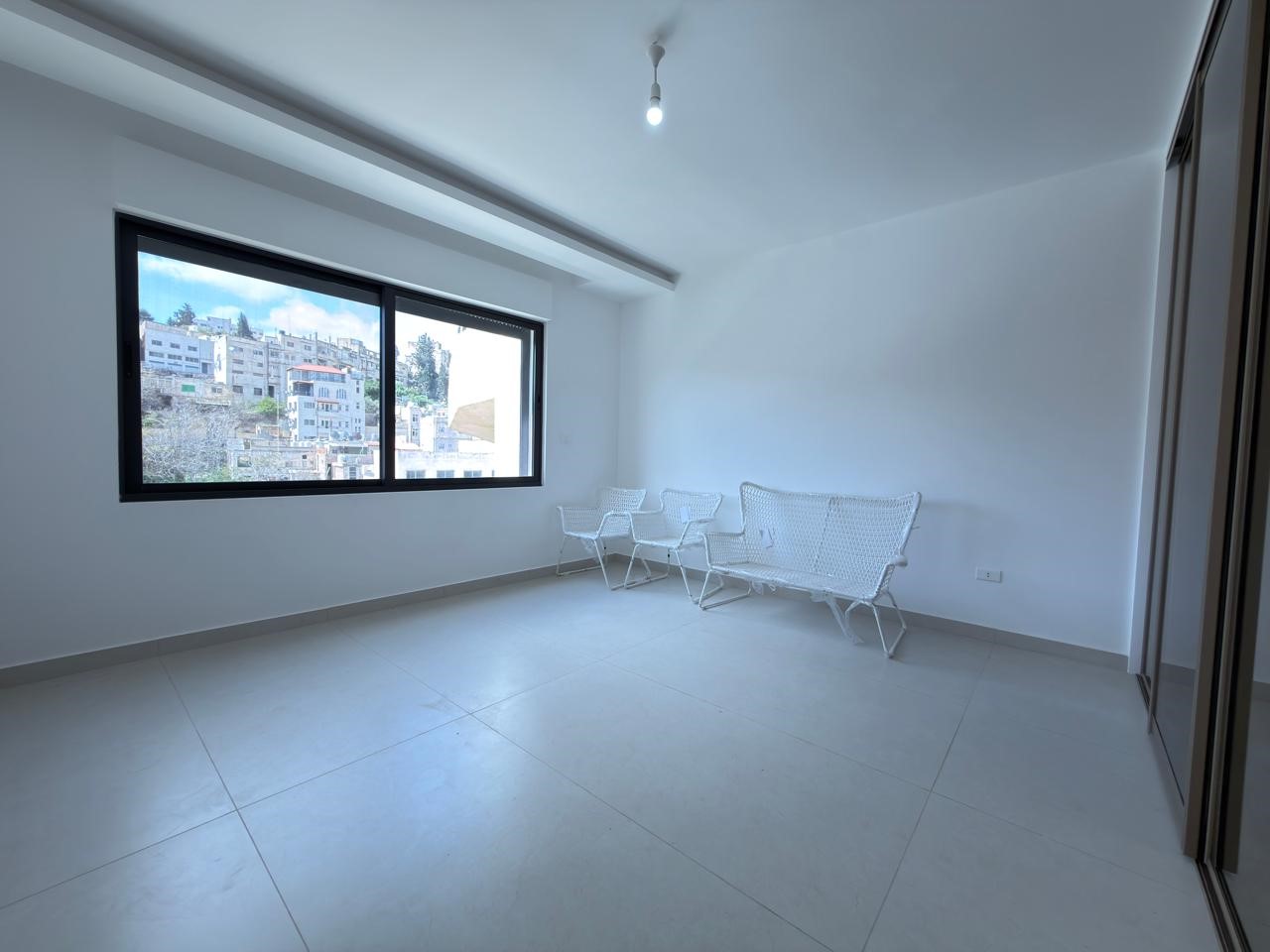 93681536-4d33-4d9d-97b6-d4f563554840 Bright minimalist room with a large horizontal window showing a hillside city view, white tiled floor, and four white metal chairs along the wall.