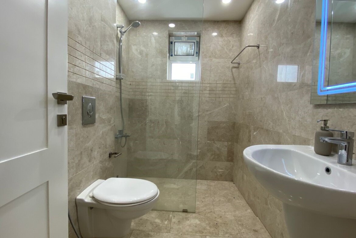 Contemporary beige tiled bathroom with a glass shower enclosure, wall-mounted toilet, and white pedestal sink.
