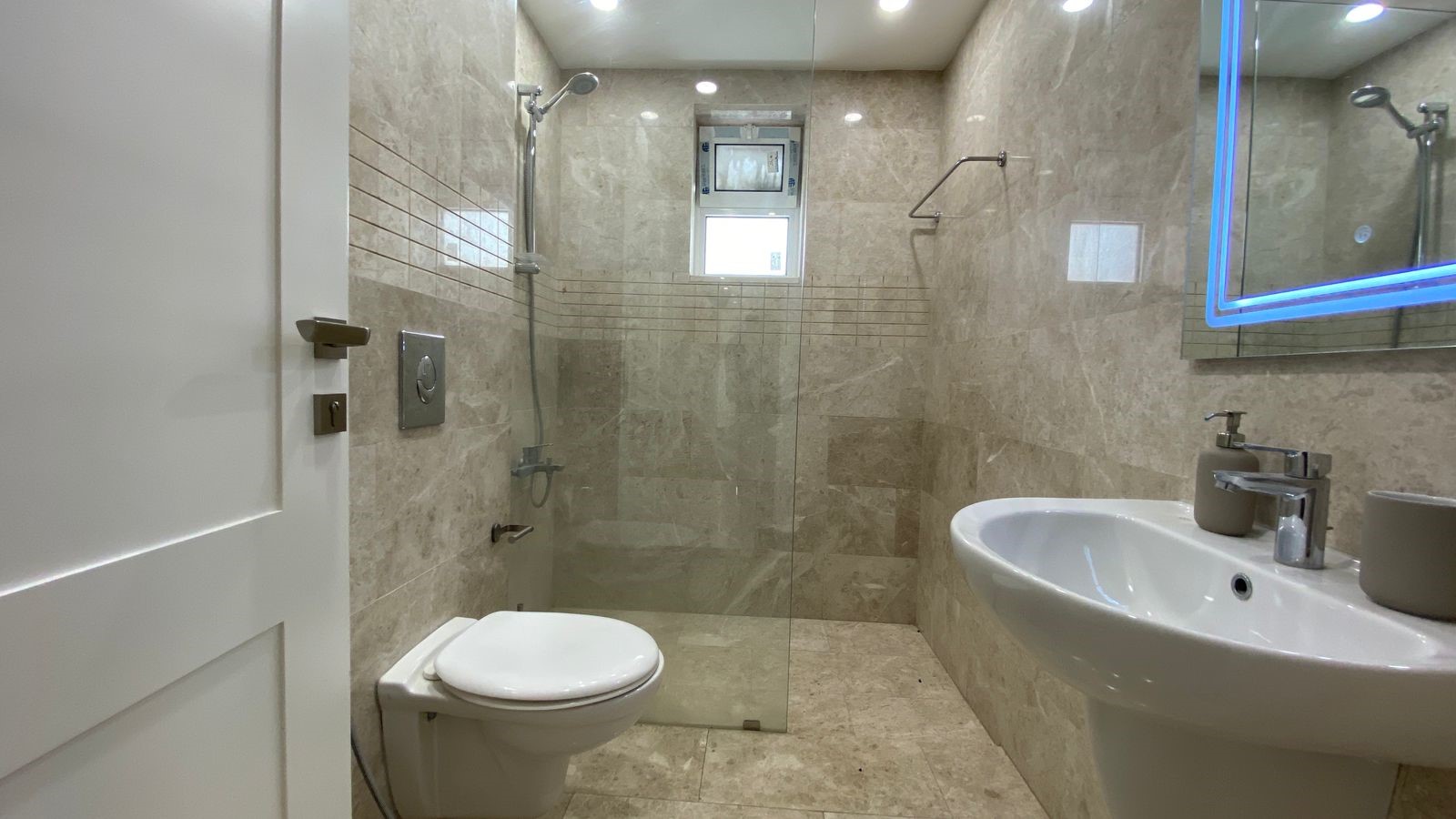 9379232e-3893-4f87-ae12-6947d4b6fdd3 Contemporary beige tiled bathroom with a glass shower enclosure, wall-mounted toilet, and white pedestal sink.