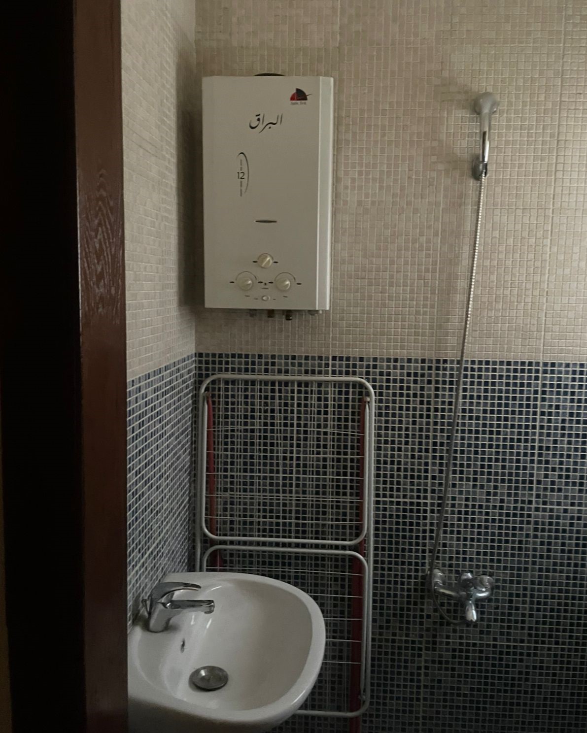 93c16611-7d03-4849-b89d-fca973b7810e Bathroom corner with a white wall-mounted boiler above a small sink and a metal towel rack on tiled walls, adjacent to a handheld shower.