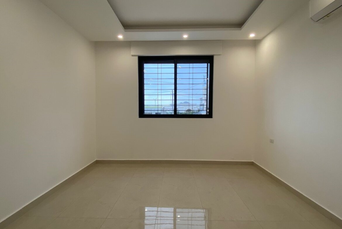 Empty, minimalist room with white walls, beige tiled floor, a centered window with metal bars, and recessed ceiling lights.