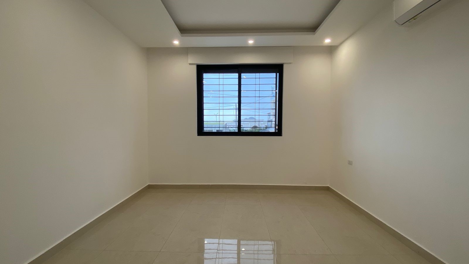 95662a3c-91c3-40bc-a826-16fb4ec17121 Empty, minimalist room with white walls, beige tiled floor, a centered window with metal bars, and recessed ceiling lights.