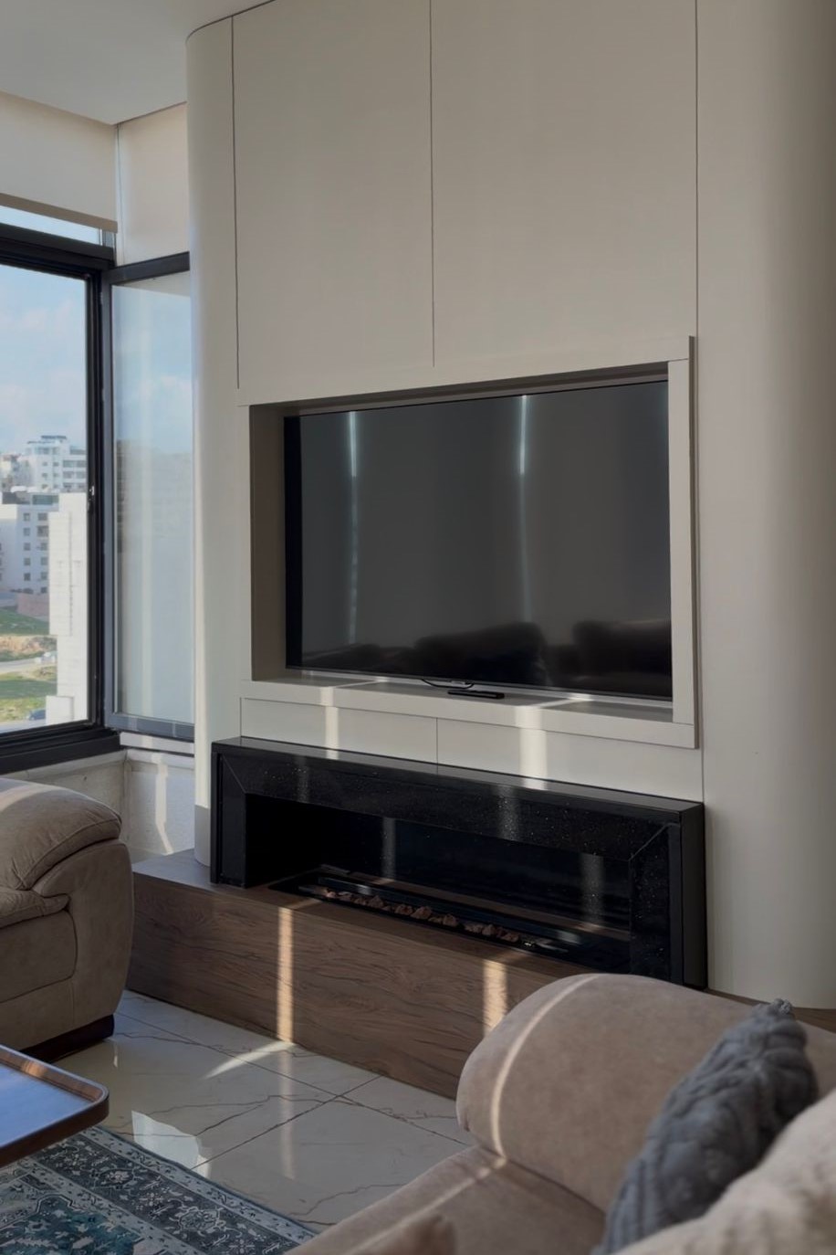 95d1c1b4-ccb4-4050-854f-d1f4c5a49553 Modern living room with a wall-mounted TV set in a built-in cabinet and a black fireplace below, next to a large window overlooking city buildings فتاة?