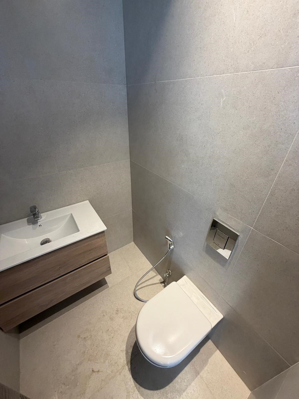 990a1c98-845a-4ea9-b37c-1eb15817b97c Modern small bathroom with a wall-mounted wooden vanity, white sink, and wall toilet with handheld bidet hose.