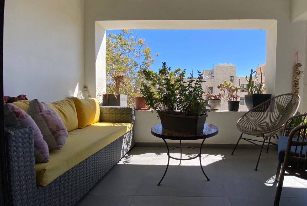 Cozy balcony seating with yellow cushions, wicker sofa, and potted plants on a round table against a bright blue sky backdrop