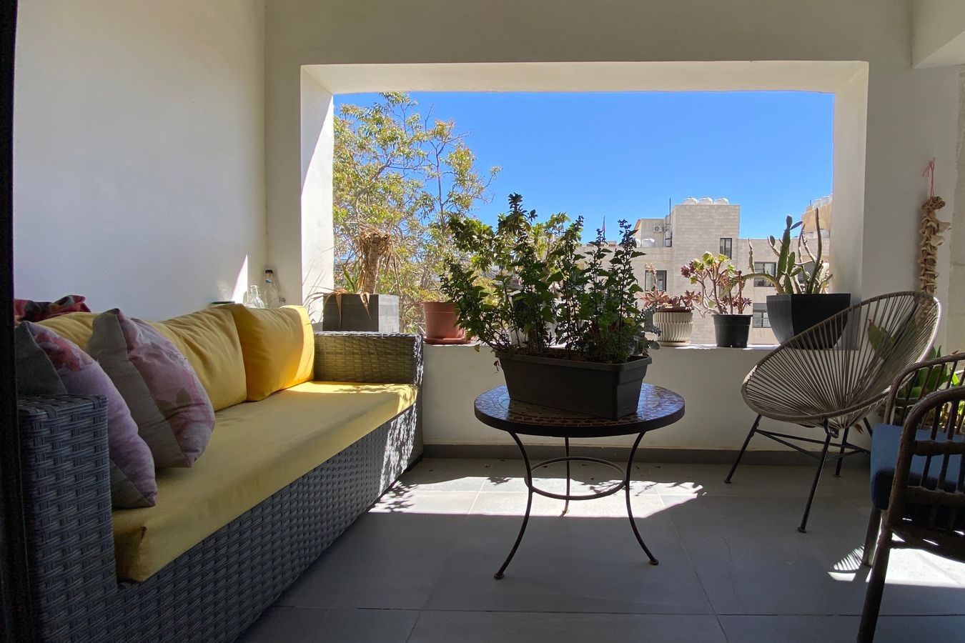 Cozy balcony seating with yellow cushions, wicker sofa, and potted plants on a round table against a bright blue sky backdrop