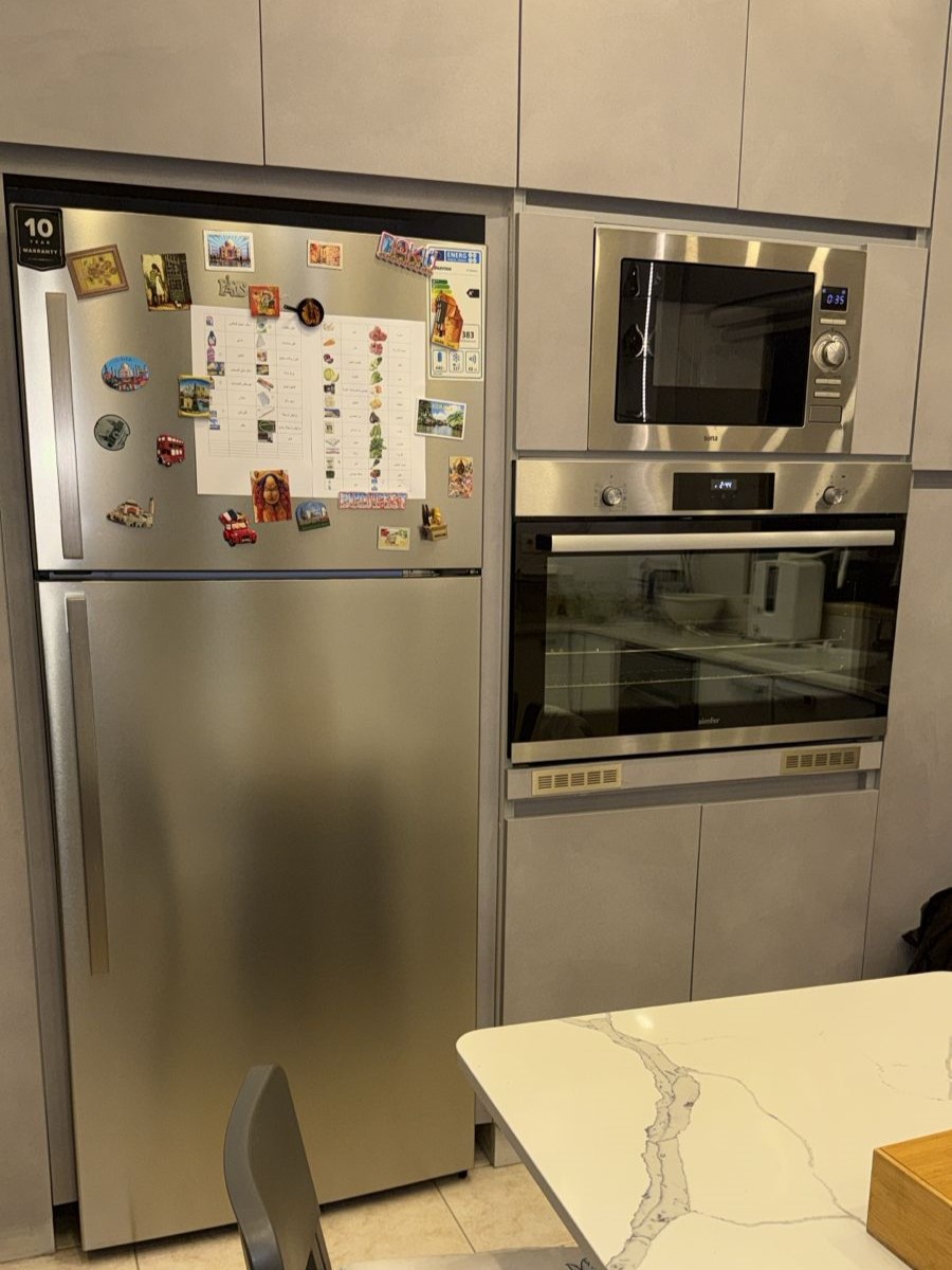 9b0e7015-e4b2-4f6c-a927-83d3d7fca16e Stainless steel fridge covered with magnets and a paper planner, next to a built‑in microwave and wall oven in a modern kitchen.