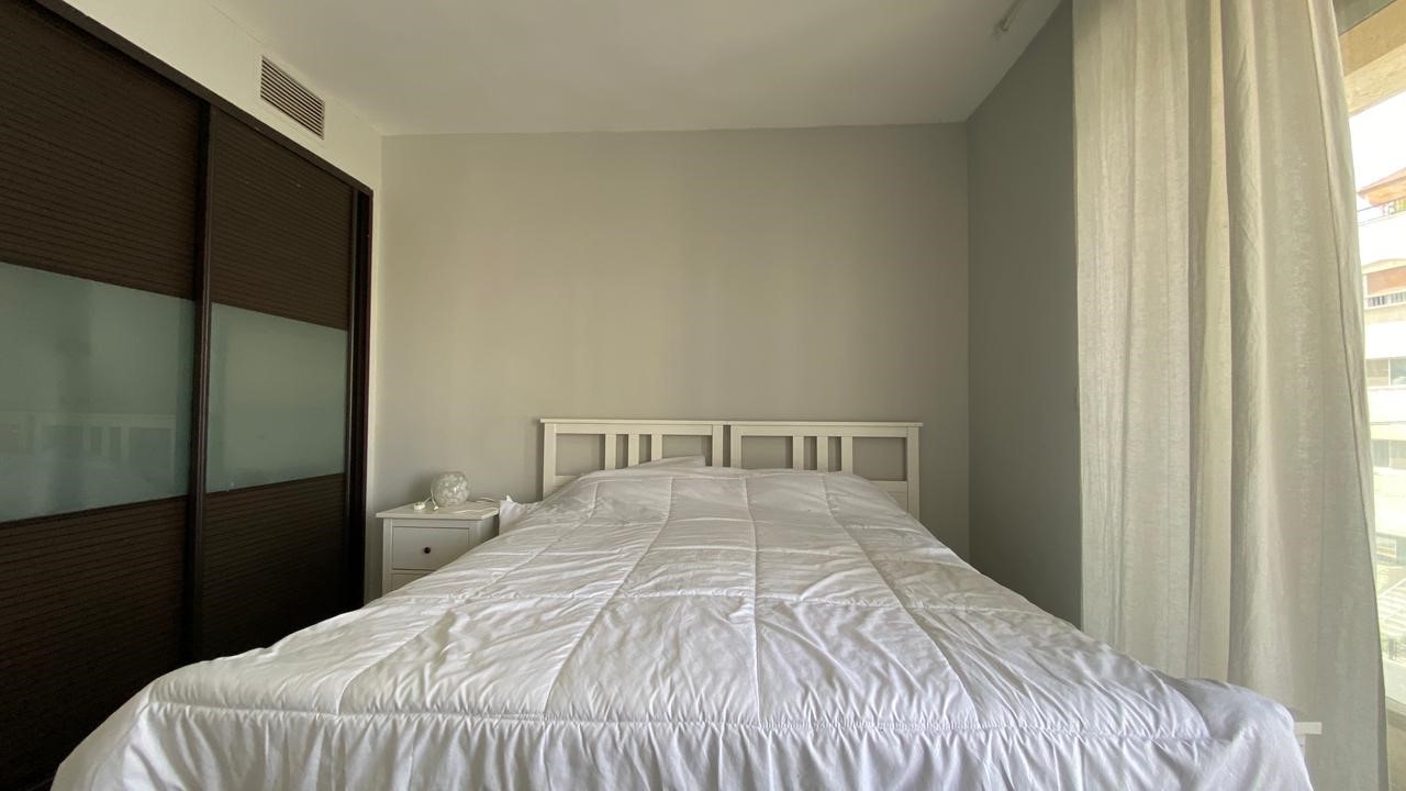 9c4aef4f-454d-4c03-a7c3-2e233527d6a6 Bright, minimal bedroom with a white metal bed frame and fluffy white bedding, flanked by a white nightstand and dark sliding wardrobe on the left. A large window with sheer curtains is on the right.