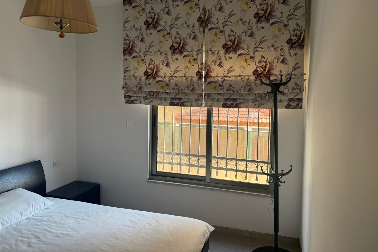 9cbbae52-43a0-44ba-9dec-d221c6d52f1a Bedroom scene: bed with white sheets and dark headboard, a black nightstand, and floral window blinds by the window. When coat rack stands nearby.
