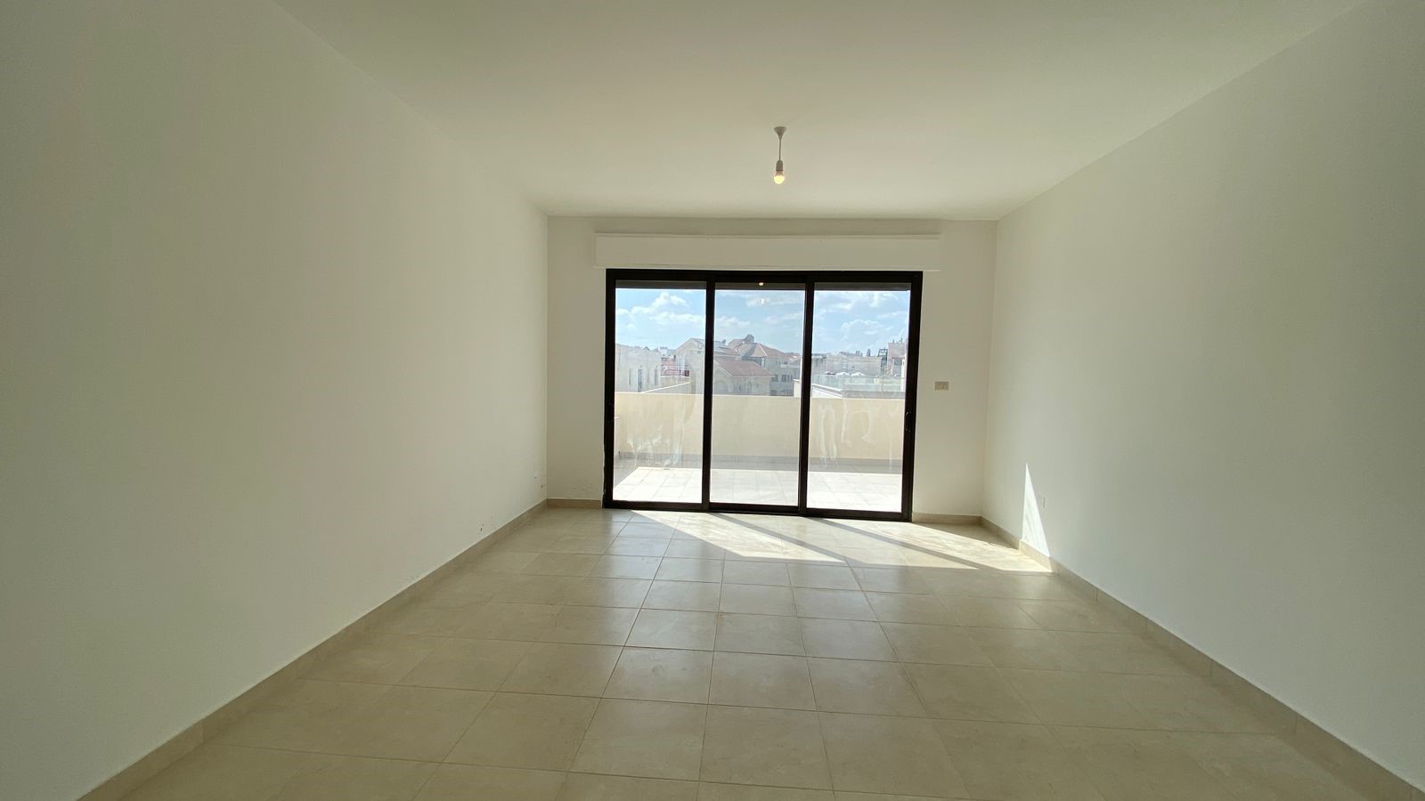 9ce0085e-c935-407a-96bf-92cf33a2b07c Empty sunlit living room with beige tiled floor and white walls, sliding glass doors to a balcony overlooking rooftops. Hugo light bulb hanging from the ceiling.