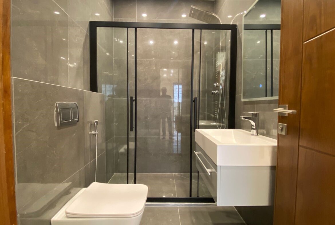 Modern bathroom with a black-framed glass shower, wall-mounted toilet, and white vanity sink on gray tiled walls and floor.