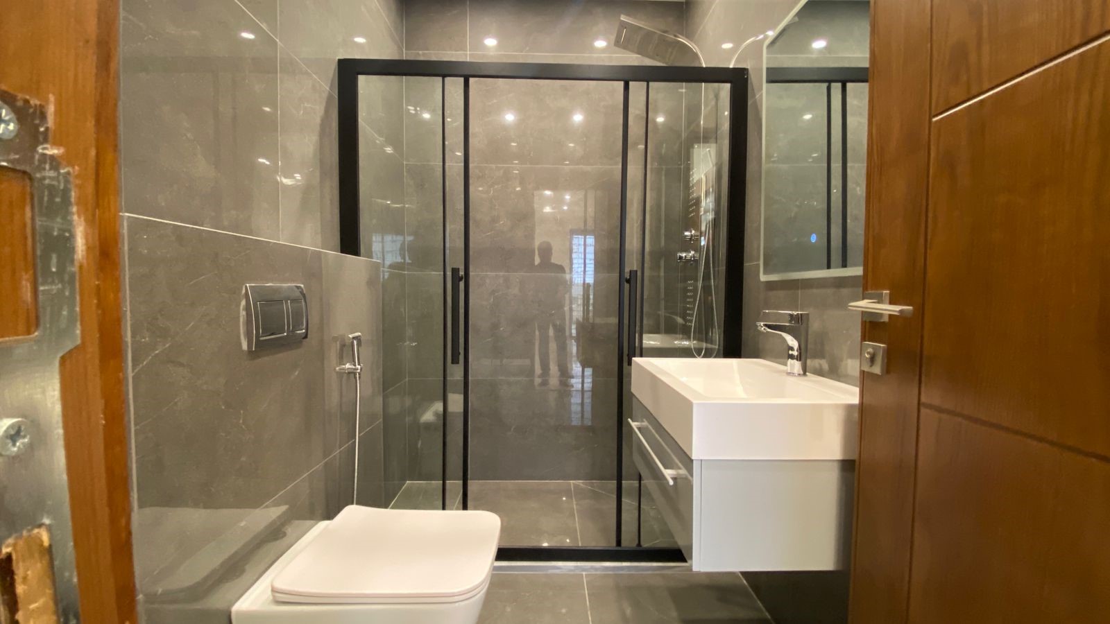 9d7d928d-36f3-46fe-94d8-8f7ce6b33984 Modern bathroom with a black-framed glass shower, wall-mounted toilet, and white vanity sink on gray tiled walls and floor.