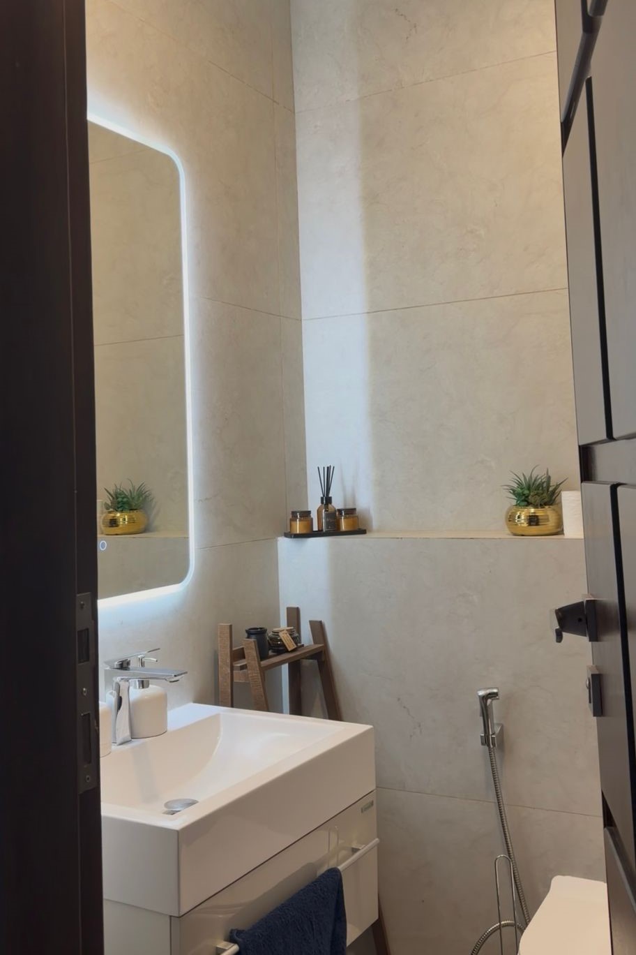 9d9a7a31-5b3d-4e30-b847-96965019655d Modern bathroom with a wall-mounted white sink, backlit mirror, and small potted plants on the shelf above the sink.