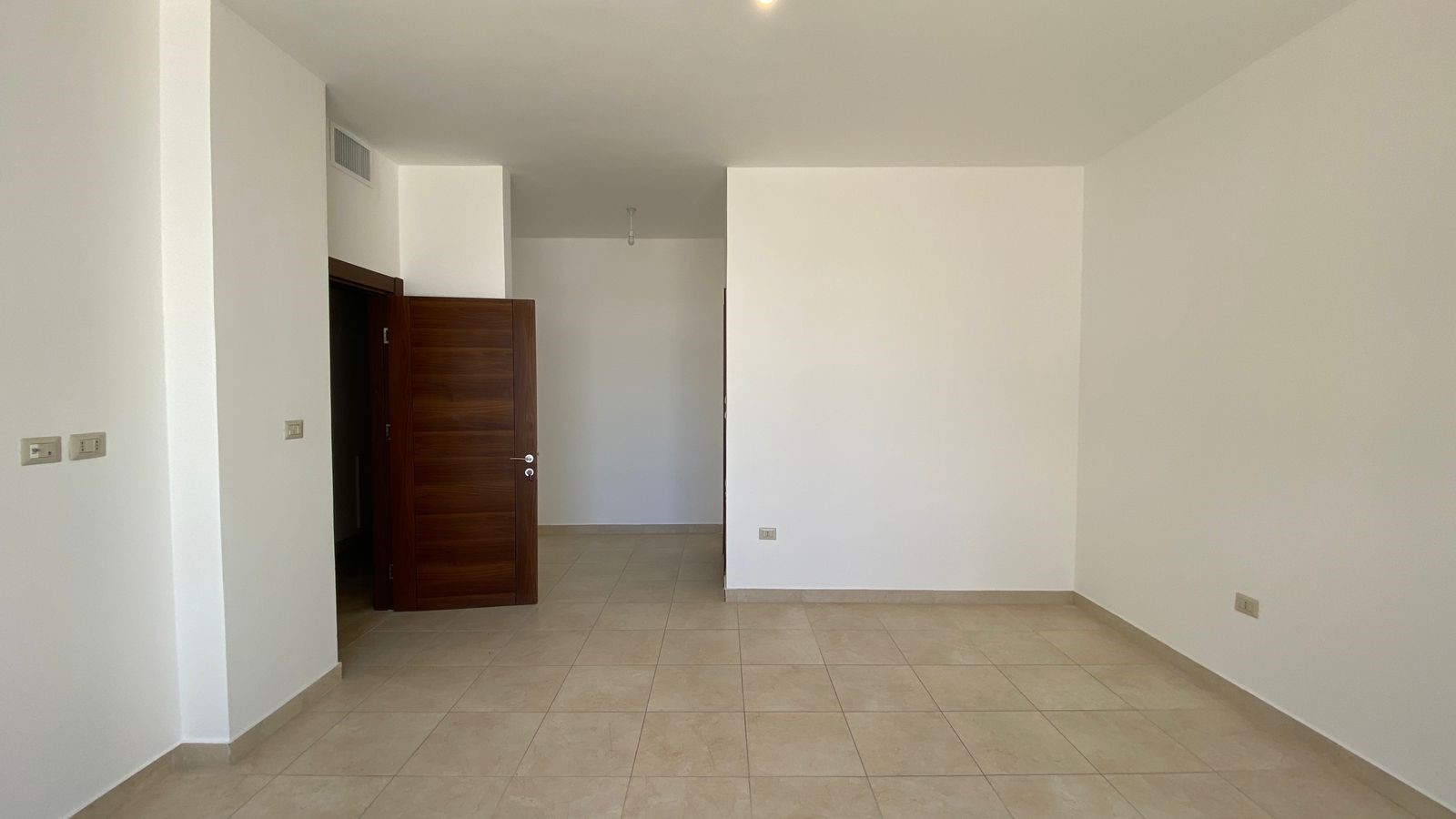 9dac508e-7521-49a3-bea0-807b9d1e972e Empty living room with beige tile floor, white walls, and a dark wood door open to a hallway.