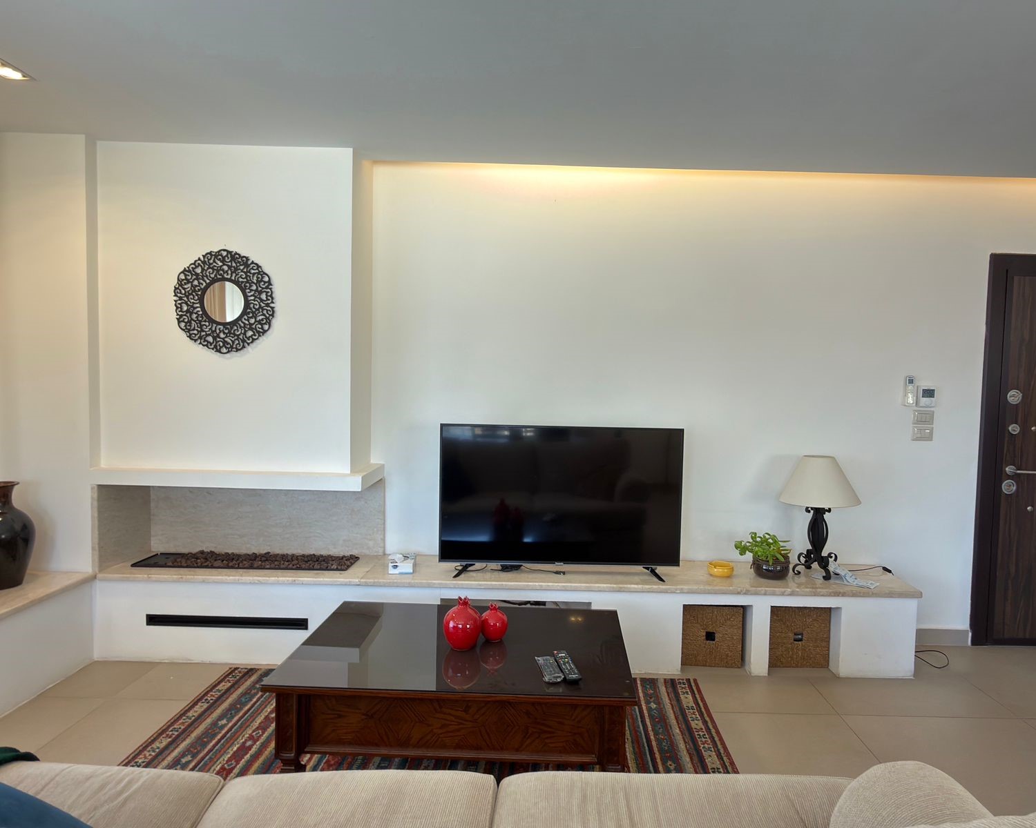 9e73a23d-b2cb-4ddc-bb80-f3141196d4e1 Living room with a flat-screen TV on a white built-in console, red decorative apples on a glass coffee table, and a patterned rug.