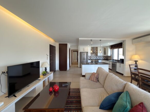 Open-plan living area with a beige sofa facing a TV on a white media console, coffee table, and a colorful rug, adjacent to a bright kitchen with an island and stainless appliances.