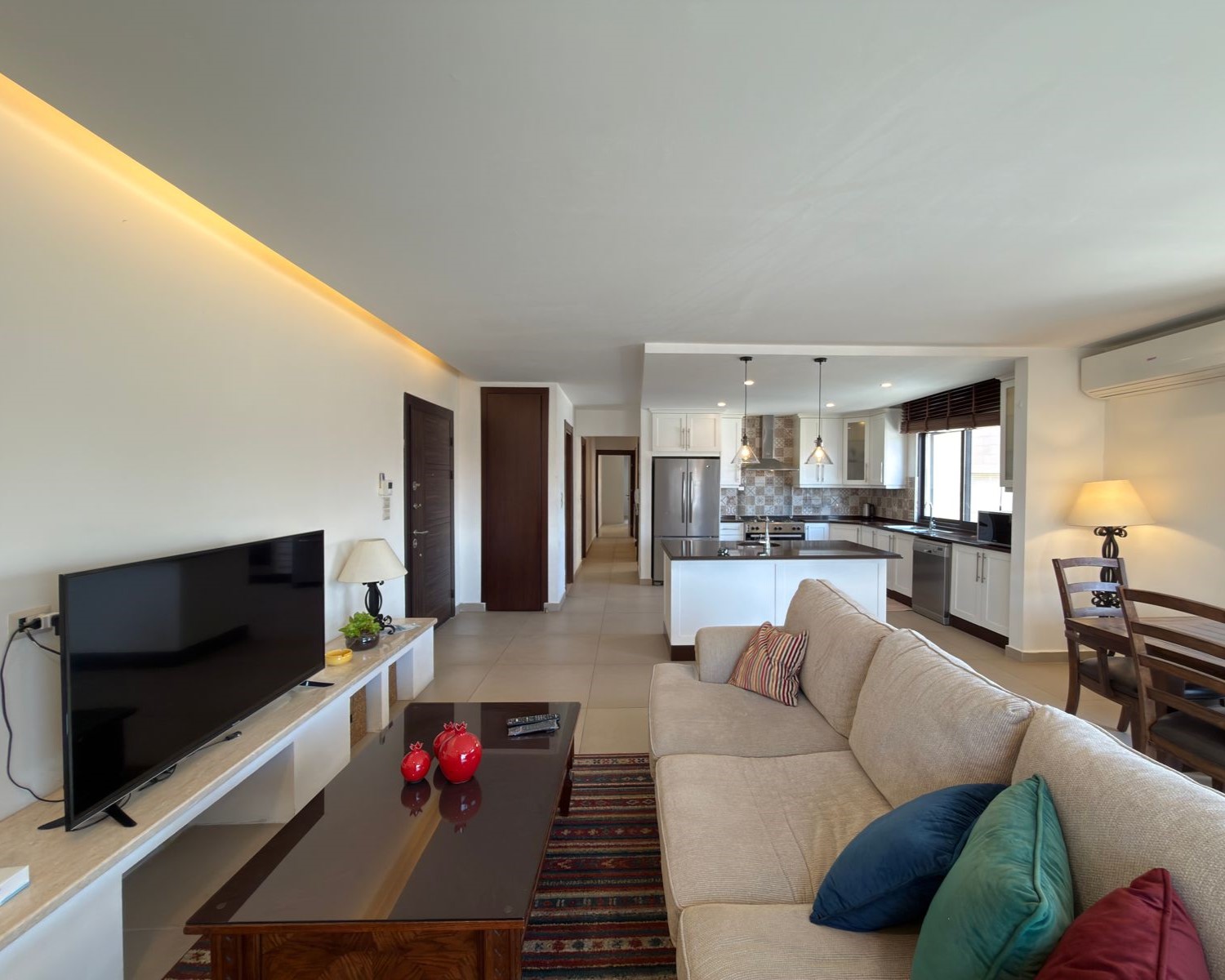 9ec54beb-1265-474a-82ba-2d1c192c0364 Open-plan living area with a beige sofa facing a TV on a white media console, coffee table, and a colorful rug, adjacent to a bright kitchen with an island and stainless appliances.