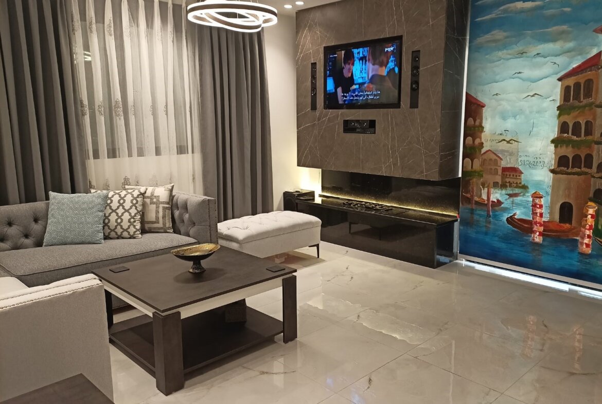 Modern living room with a wall-mounted TV above a black marble fireplace, a grey sectional sofa with decorative cushions, and a colorful seaside mural on the right wall.