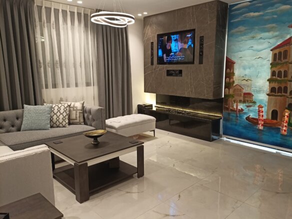 Modern living room with a wall-mounted TV above a black marble fireplace, a grey sectional sofa with decorative cushions, and a colorful seaside mural on the right wall.