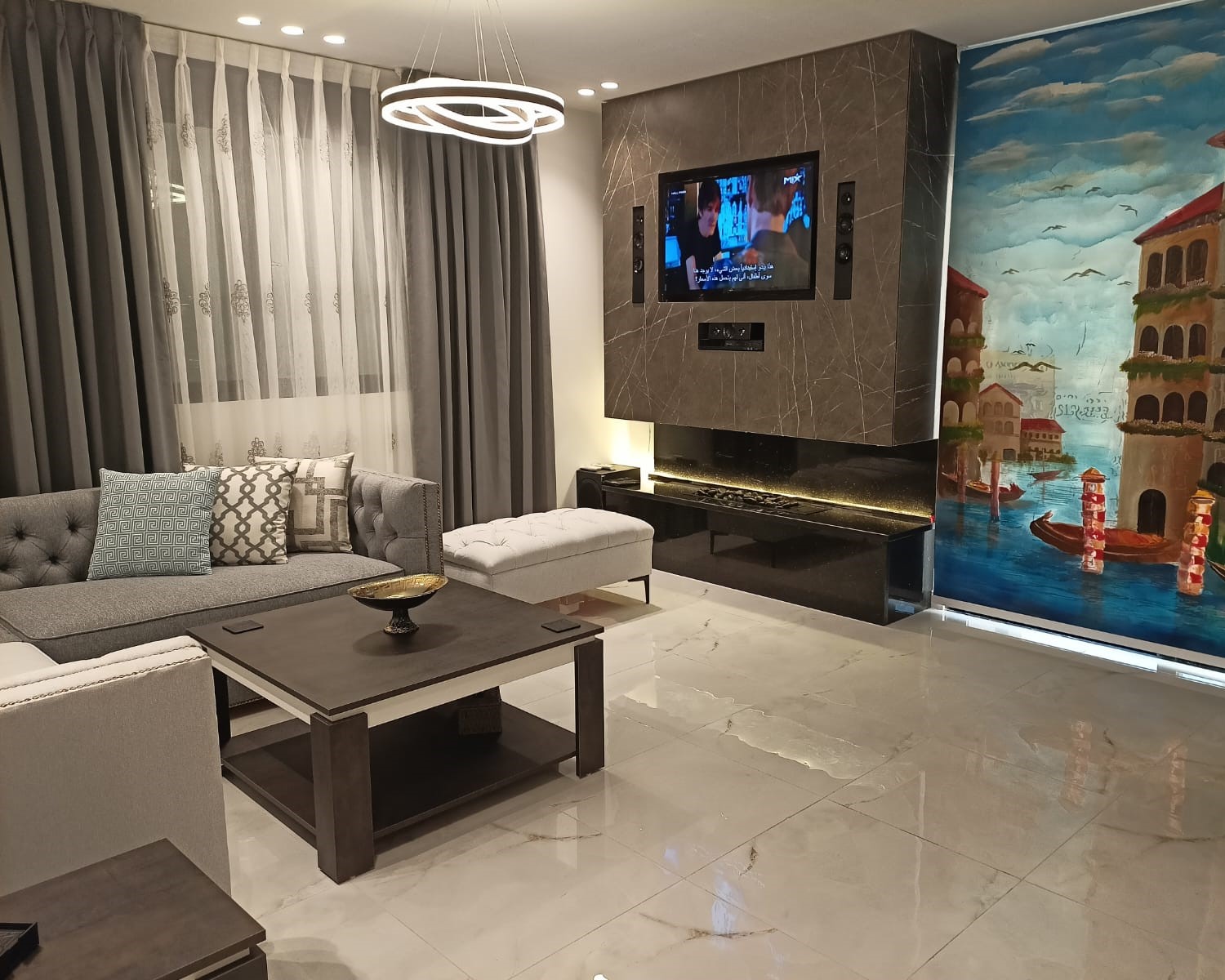 9fa26677-6357-4ce2-92d8-aec3c26cbcbc Modern living room with a wall-mounted TV above a black marble fireplace, a grey sectional sofa with decorative cushions, and a colorful seaside mural on the right wall.