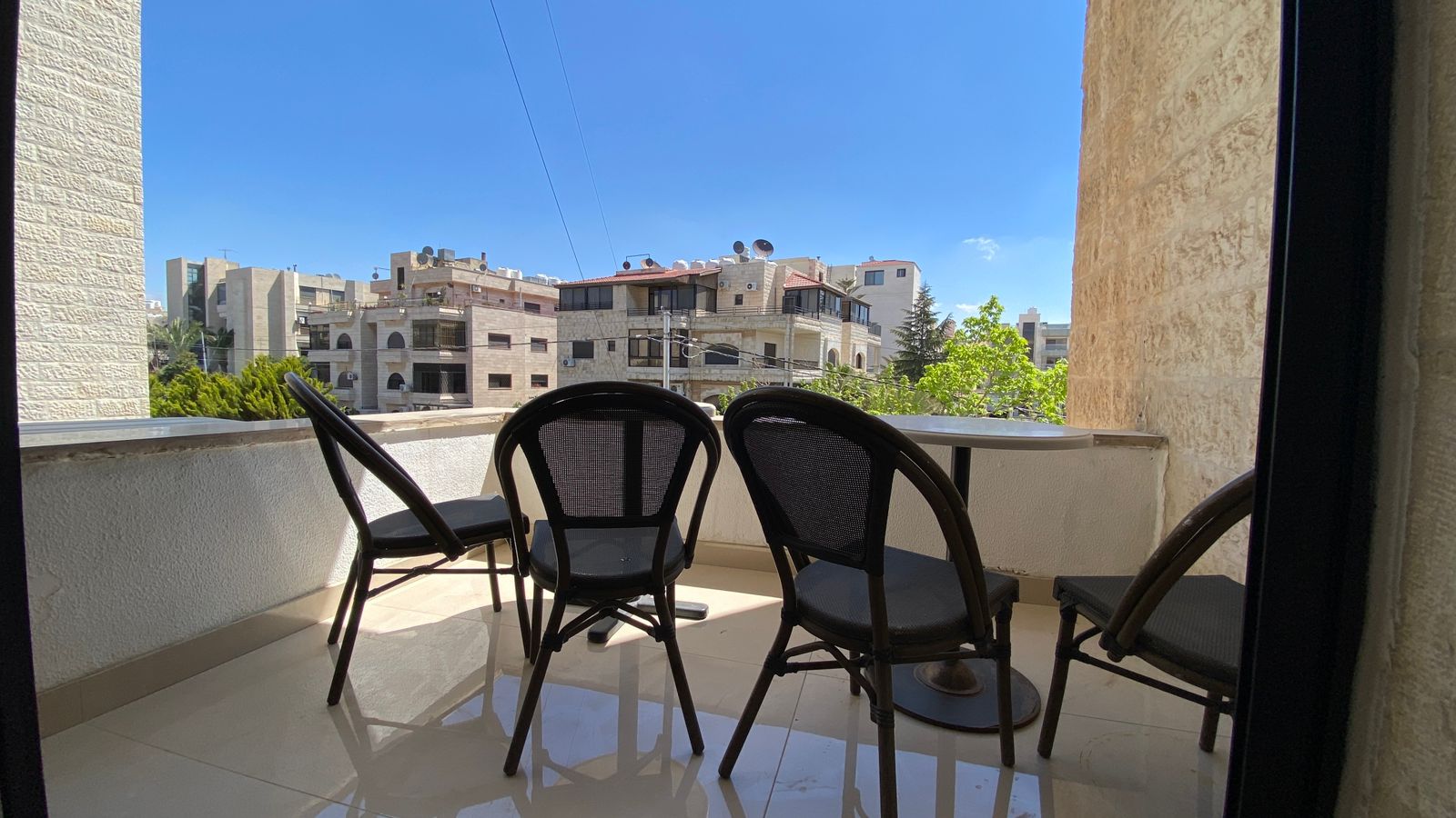 WhatsApp Image 2026-04-21 at 122948 PM 3 Balcony with four black metal chairs around a round glass table, overlooking a cityscape under a clear blue sky.