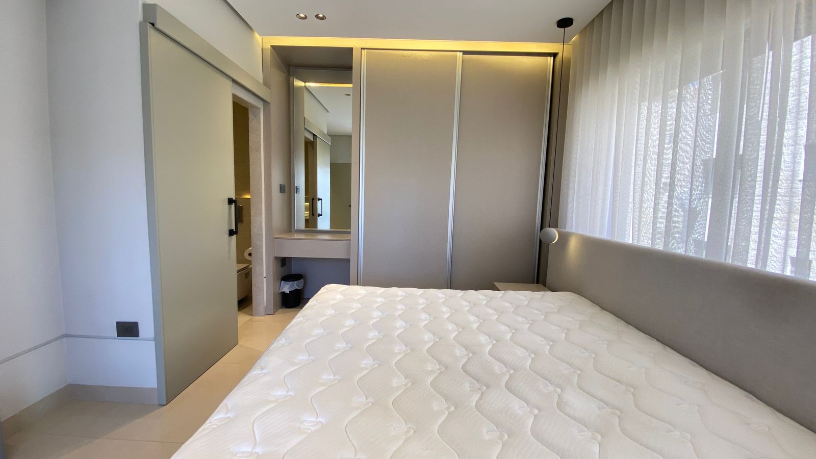 WhatsApp Image 2026-04-21 at 122949 PM 2 Modern bedroom with a large bed, quilted white mattress, sheer curtains, and a large sliding wardrobe; an open doorway reveals a small bathroom with a vanity mirror on the left.