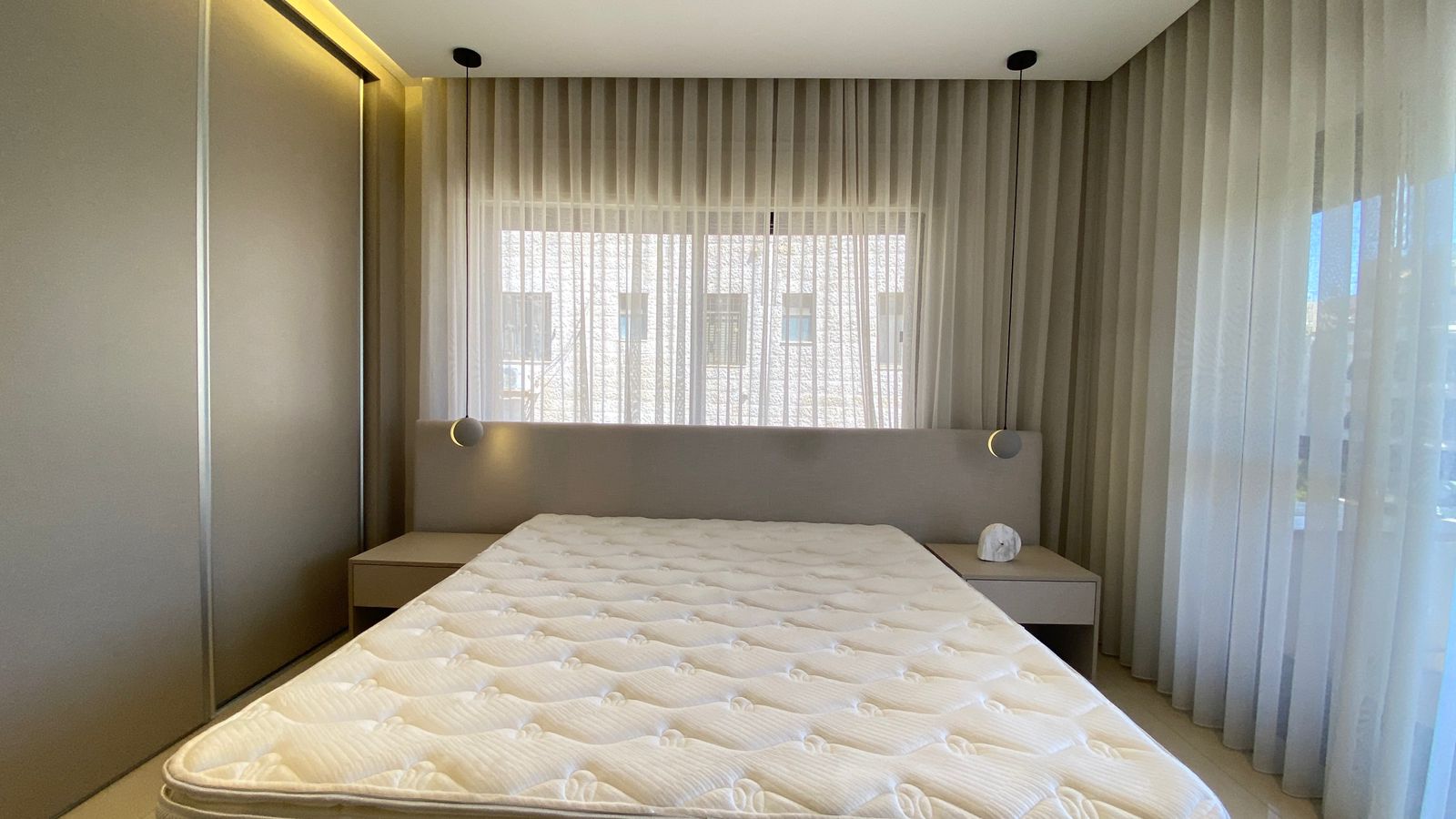 WhatsApp Image 2026-04-21 at 122949 PM 3 Minimalist bedroom with a large bed, beige headboard, two nightstands, and sheer curtains filtering sunlight.