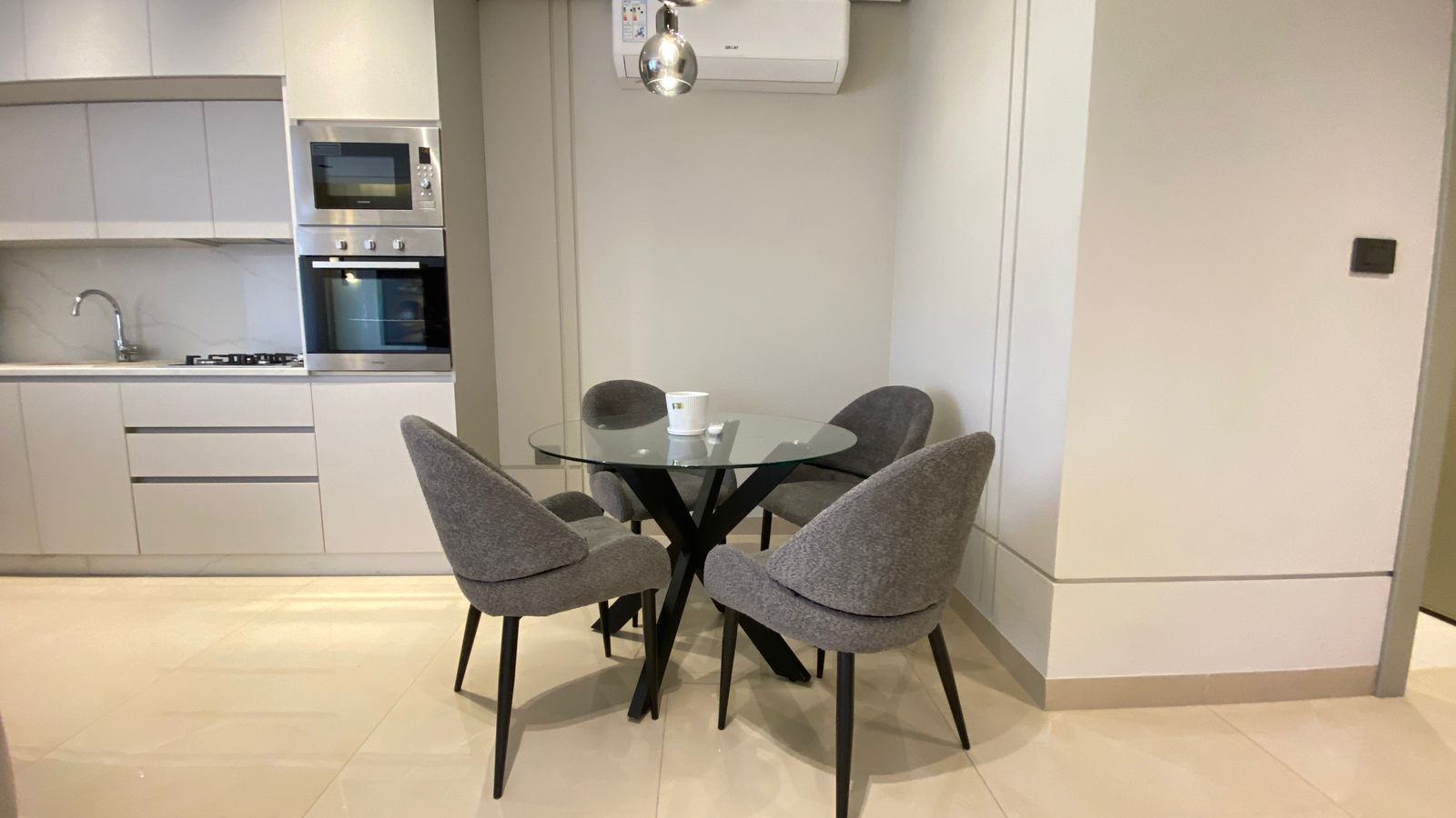 WhatsApp Image 2026-04-21 at 122950 PM 3 Modern kitchen with a round glass dining table and four gray upholstered chairs; built-in oven and microwave in white cabinets to the left.