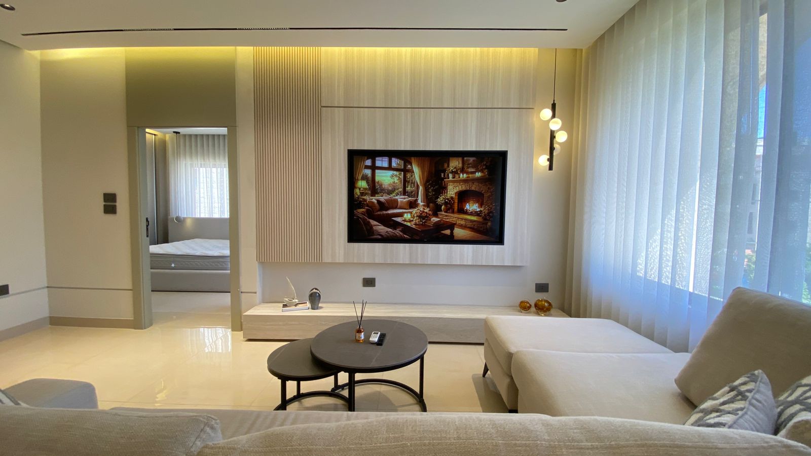 WhatsApp Image 2026-04-21 at 122950 PM 4 Contemporary living room with a wall-mounted TV framed by light wood panels, a white sectional sofa, and sheer-curtain windows.
