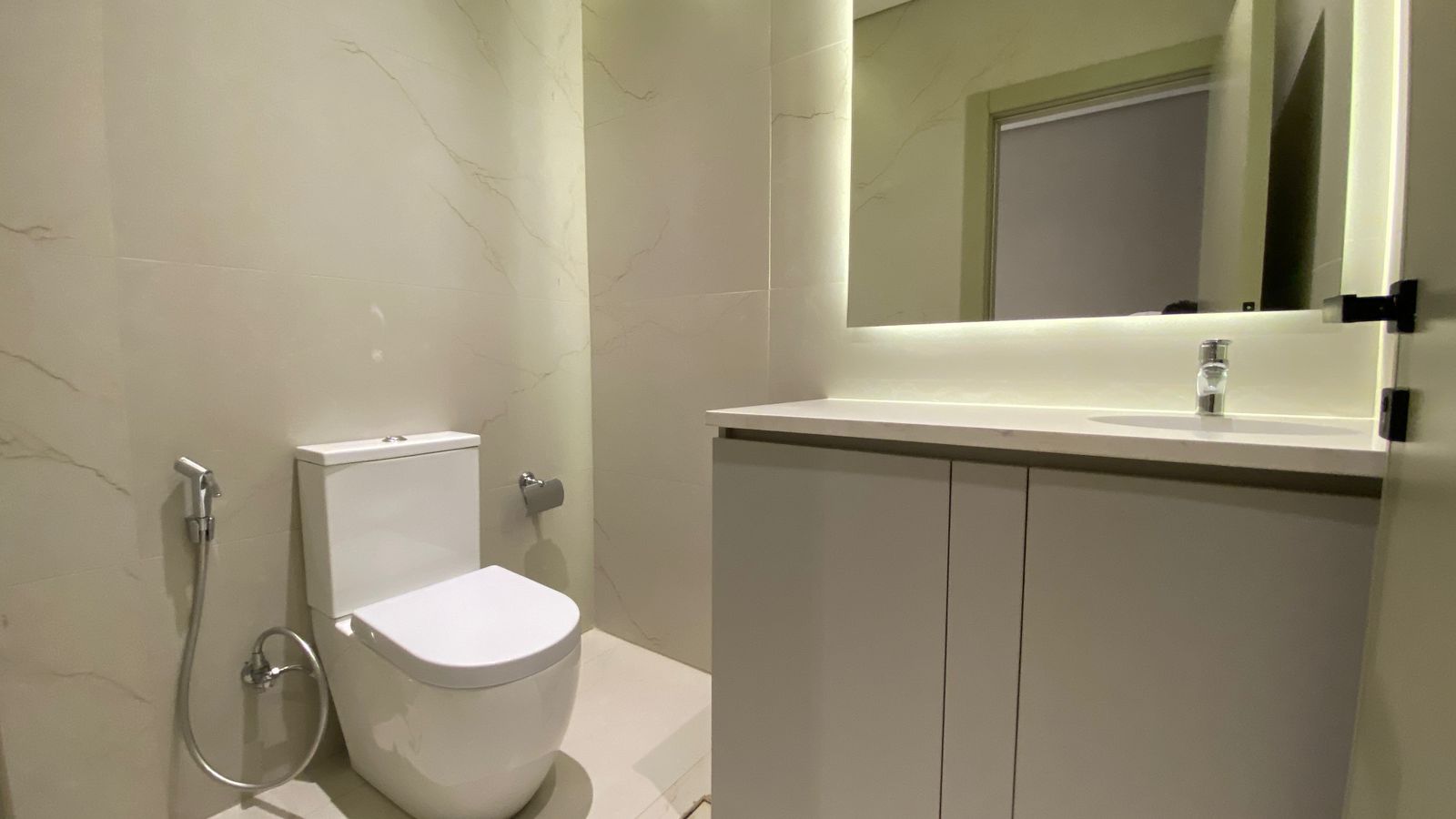WhatsApp Image 2026-04-21 at 122950 PM Modern bathroom with a white toilet, bidet spray, and a gray vanity under a backlit mirror.