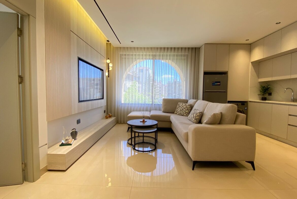 Bright modern living room with an L-shaped beige sofa, round nesting coffee tables, and a wall-mounted TV under a wood-accent wall. The large arched window with sheer curtains lets in natural light.