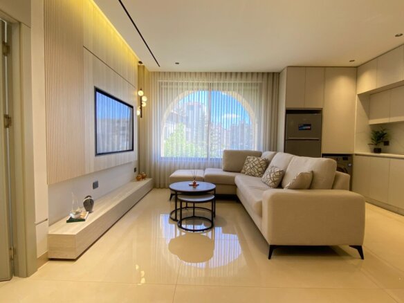 Bright modern living room with an L-shaped beige sofa, round nesting coffee tables, and a wall-mounted TV under a wood-accent wall. The large arched window with sheer curtains lets in natural light.