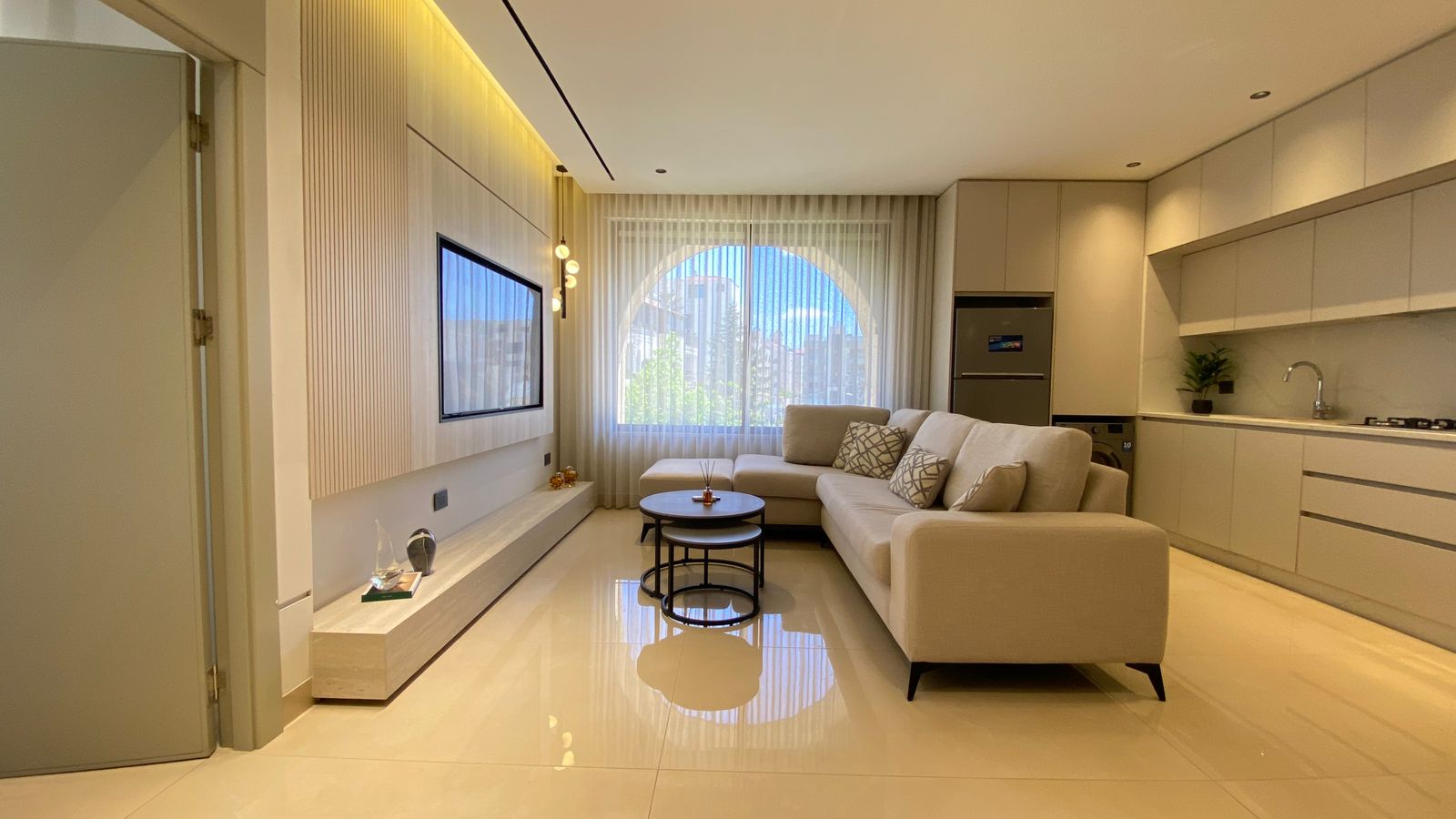 WhatsApp Image 2026-04-21 at 122951 PM Bright modern living room with an L-shaped beige sofa, round nesting coffee tables, and a wall-mounted TV under a wood-accent wall. The large arched window with sheer curtains lets in natural light.