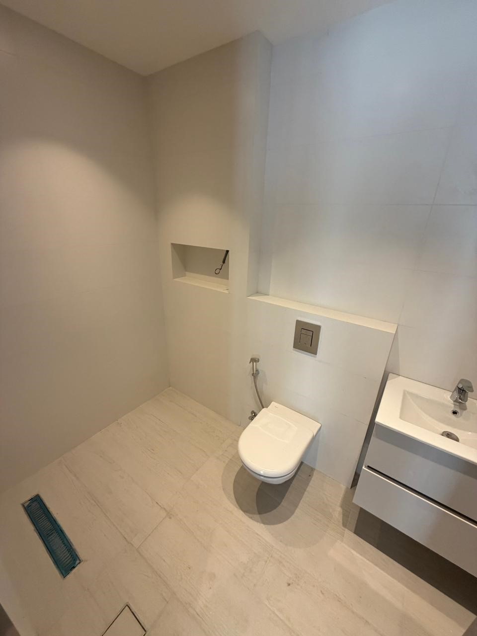 a15ad33e-31da-45d3-a60f-6800ae540c15 Modern minimalist bathroom with a wall-mounted toilet, white vanity, and a recessed shower niche on tiled walls and floor.