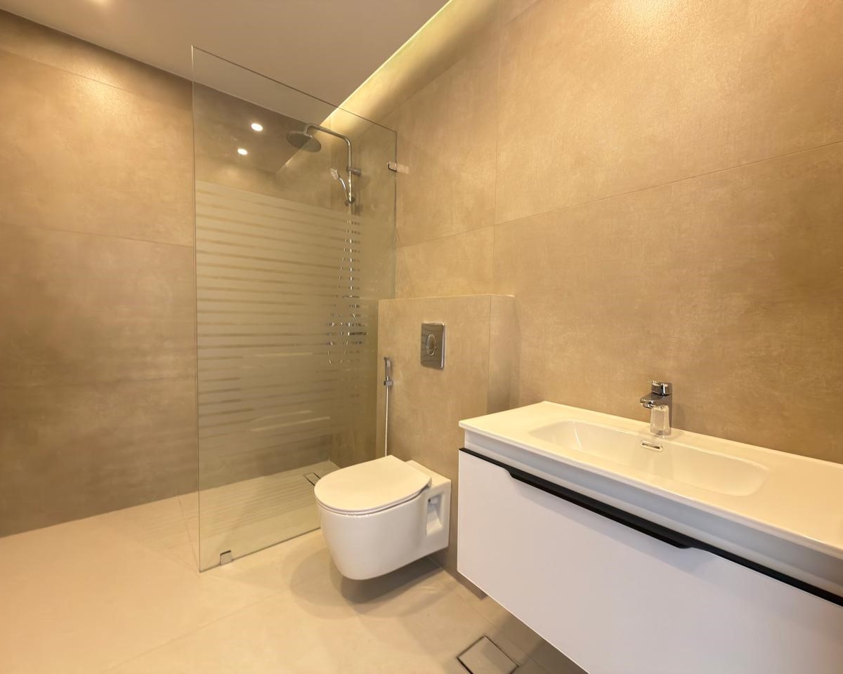a19e67a1-0103-4de0-bfef-4f821b08a335 Modern beige-tiled bathroom with glass-enclosed shower, wall-mounted toilet, and white vanity with sink.