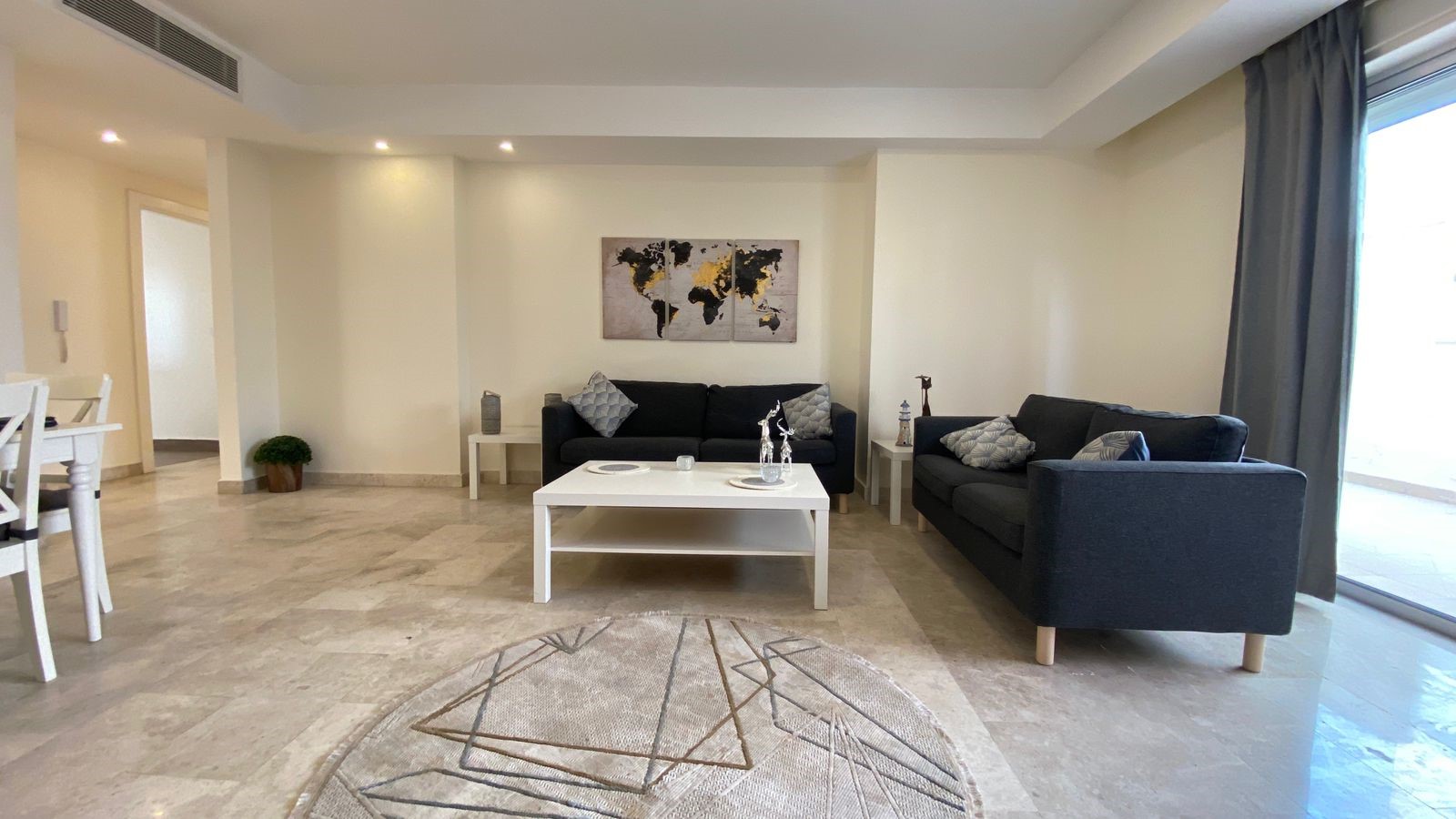 a1d2fbae-c9ea-4e9c-aaa5-81147bc37ed0 Modern living room with two dark gray sofas, a white coffee table, world map wall art, and a large window with gray curtains.