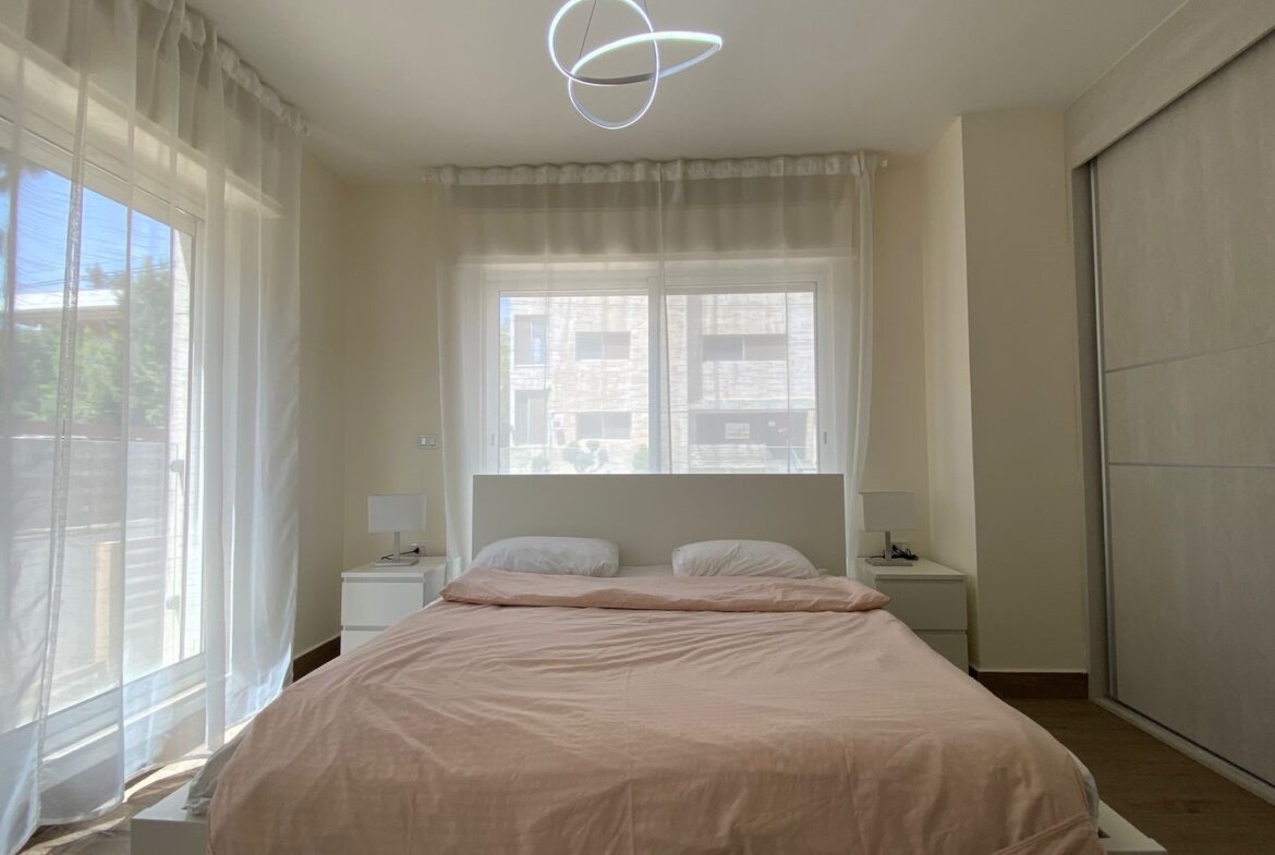 Bright minimalist bedroom with a pink bedspread, white headboard, two white pillows, and nightstands by the windows.