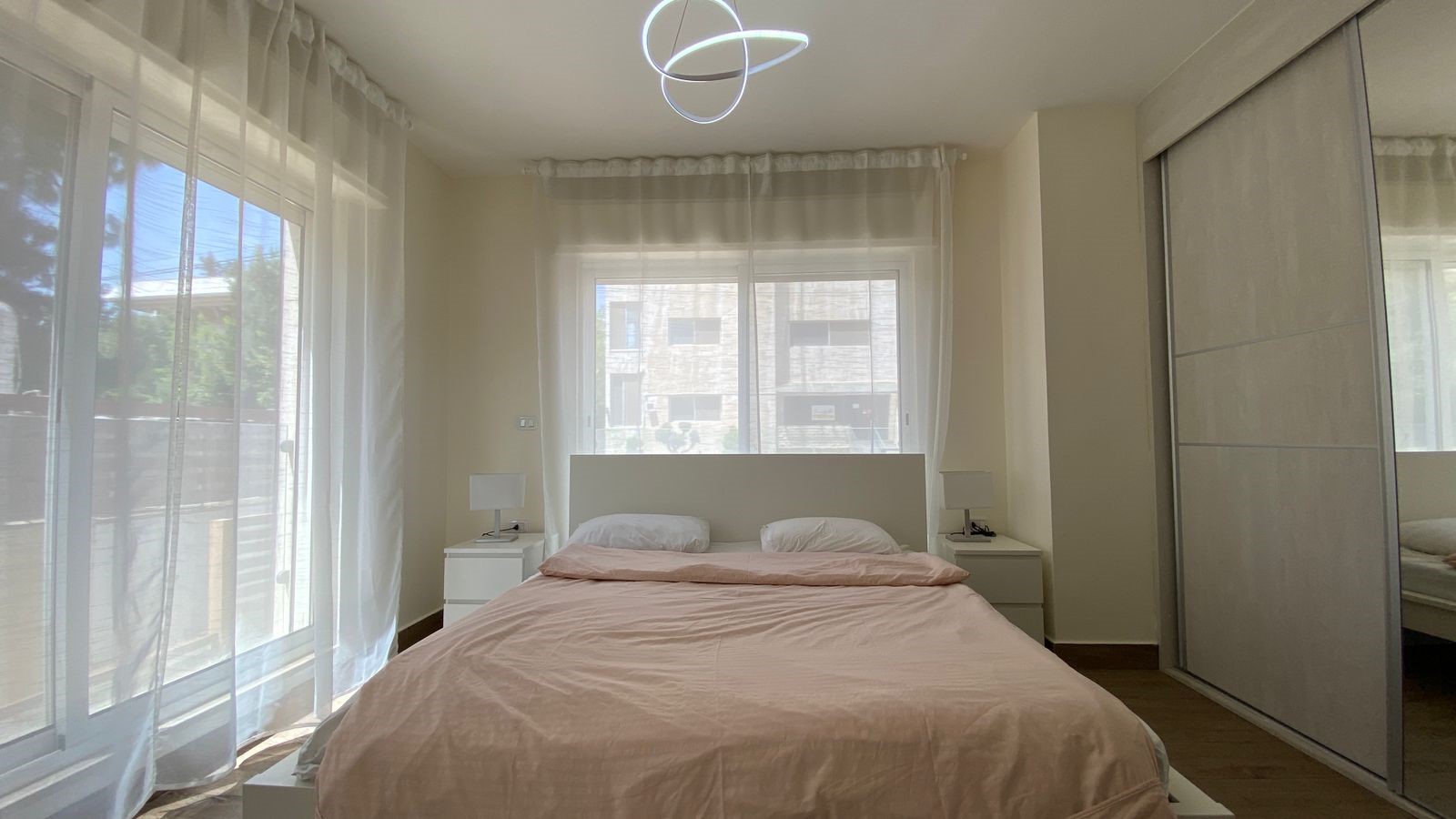 a1ffefd0-0474-4e89-a86d-6edf692da0fc Bright minimalist bedroom with a pink bedspread, white headboard, two white pillows, and nightstands by the windows.