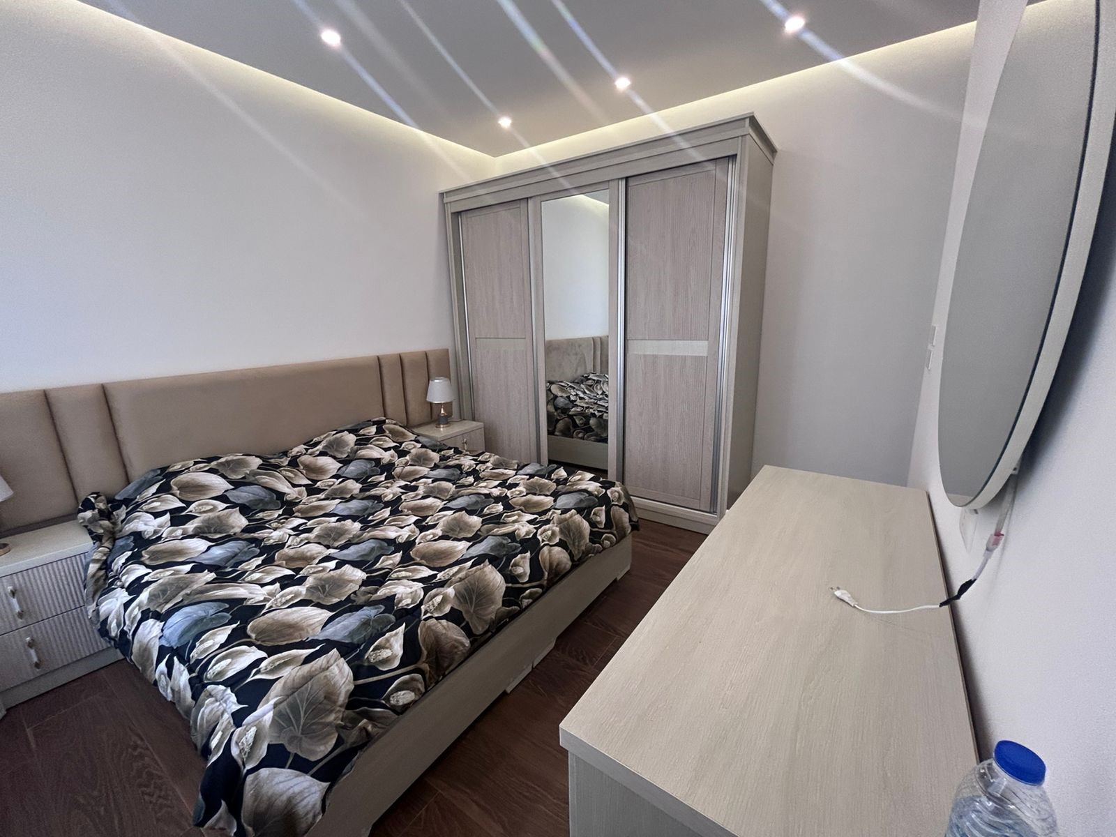 a2115108-d401-47f4-b237-3cc531ec822b Modern beige bedroom with a double bed, floral duvet, two matching nightstands, and a tall wardrobe with mirrored doors.