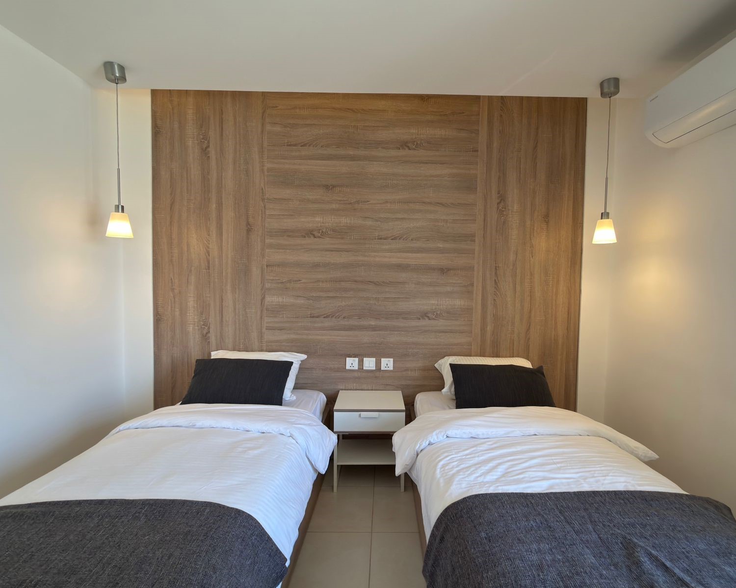 a44fa5f1-672b-451a-ac9c-8b9bbeee615b Twin bedroom with a wood-paneled headboard wall, two single beds in white linens, and a shared nightstand between them.