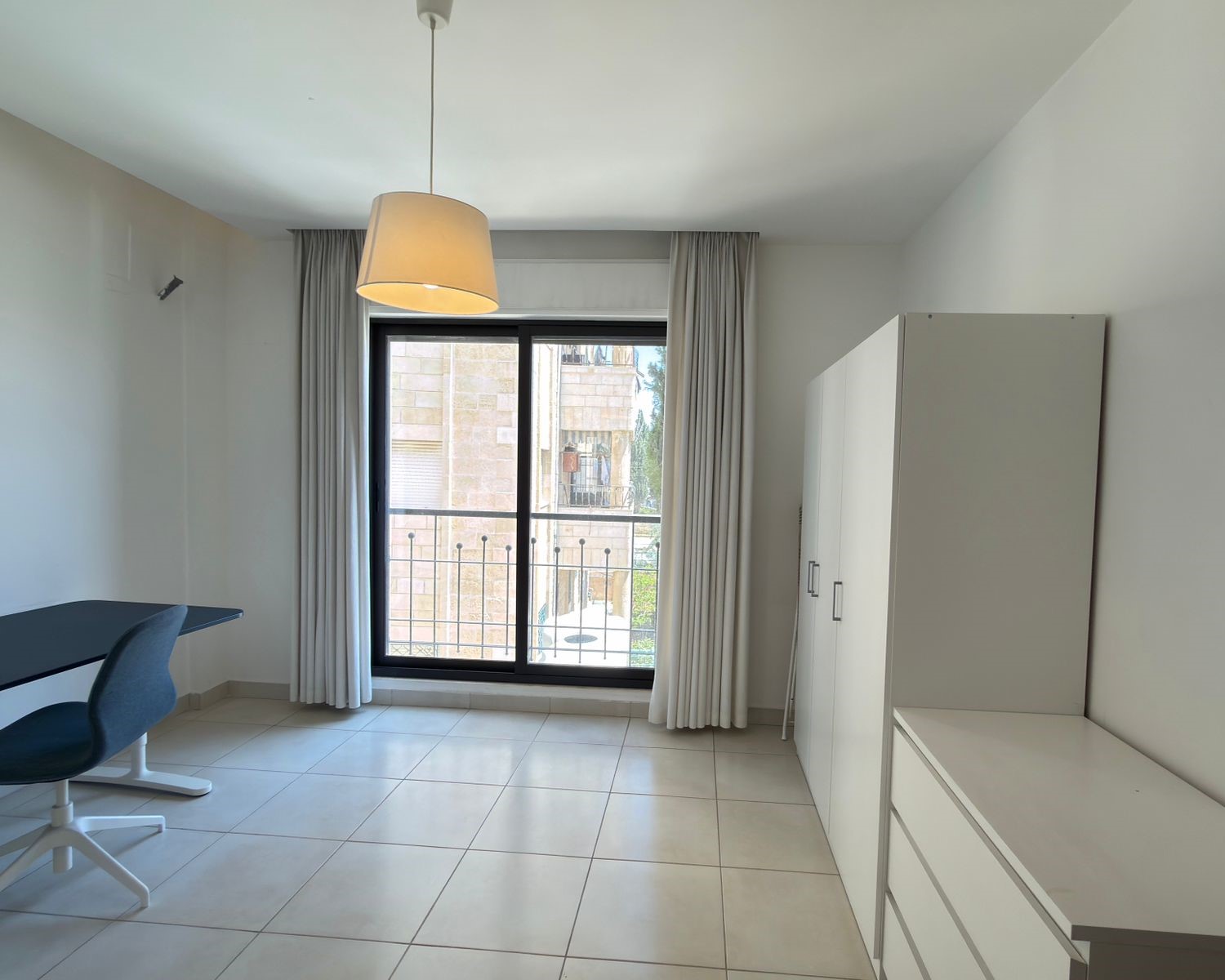 a4aa066a-2381-4308-8c5c-88031fedfc6d Bright minimalist room with a black-framed window and balcony railing, white furniture, desk and blue chair, pendant light overhead.