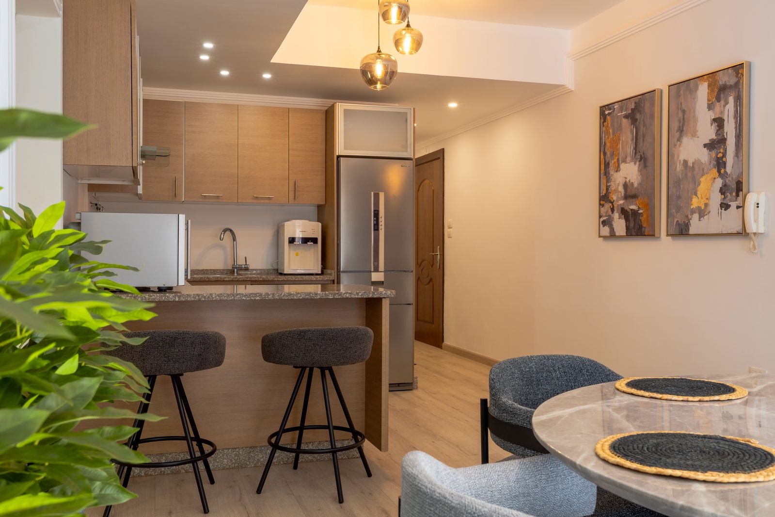 a55e6c47-ab54-443c-be46-0f481fe3a6b6 Open-plan kitchen with a granite island, wooden cabinets, and a stainless steel refrigerator; dining chairs at the island and a round marble table nearby.