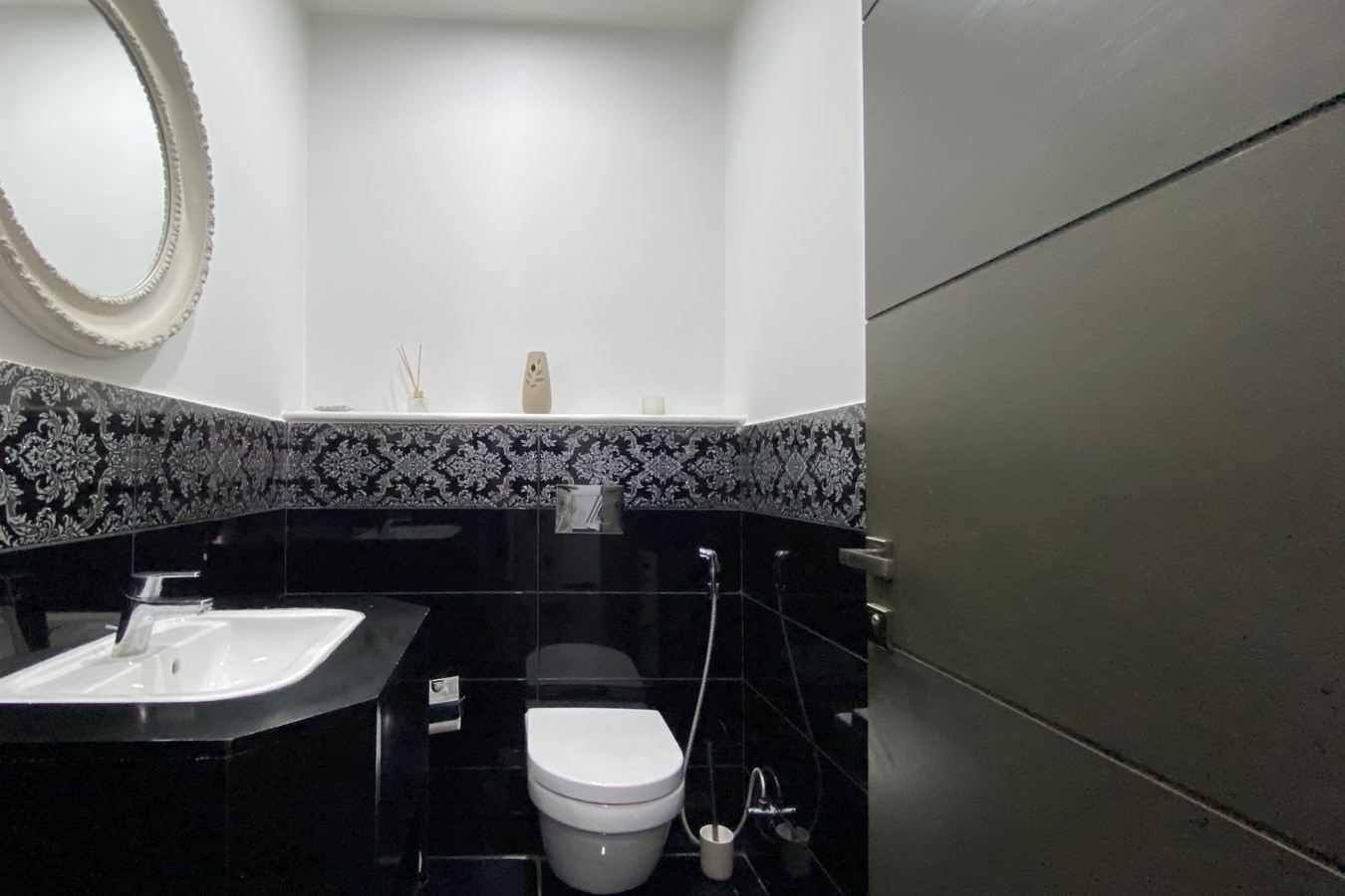 a5add811-3e12-4a6c-a476-87b15c9a707c White bathroom with black tile midsection, ornate round mirror, and white sink on black cabinet surface side by side with a toilet and bidet hose.