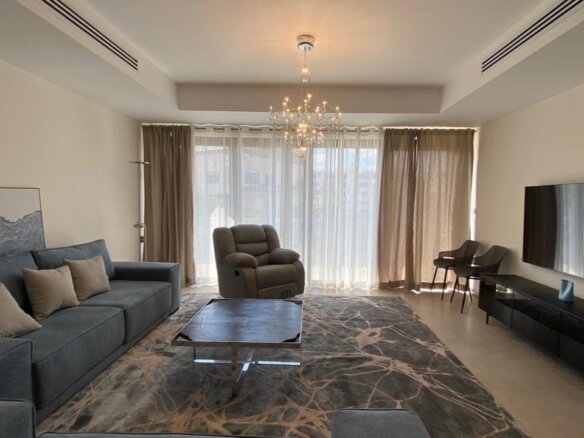 Bright modern living room with a blue sofa, beige cushions, and a gray recliner facing a wall-mounted TV; large windows with sheer curtains fill the space with natural light.